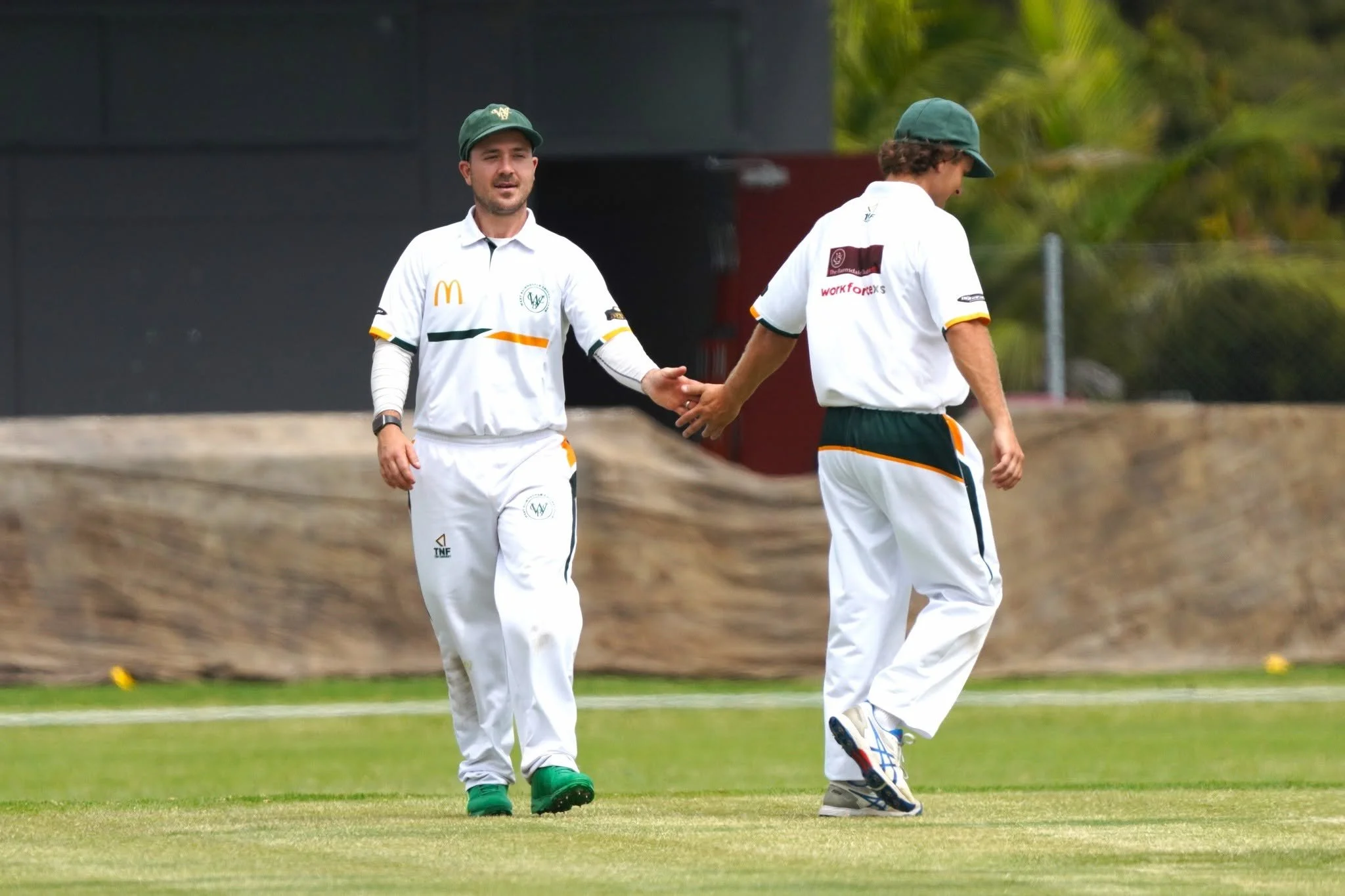 Two male cricket players wearing white uniforms and green caps, walking on a cricket field, with one player holding the other's hand.