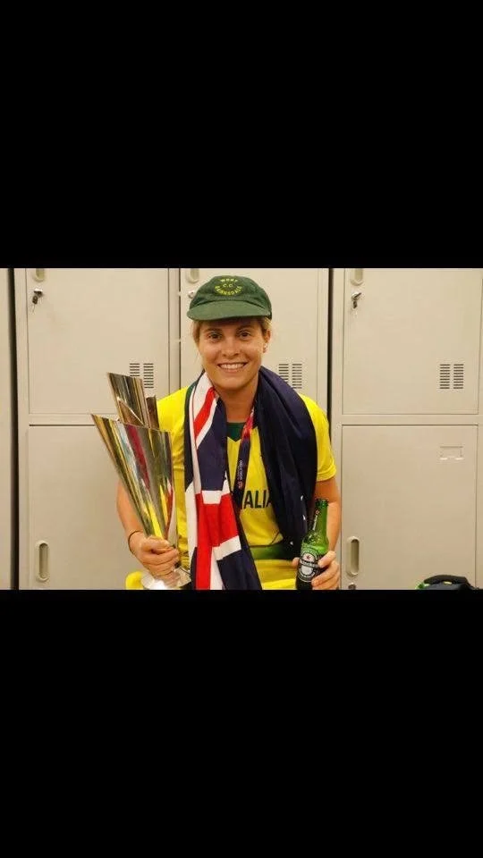 A woman dressed in a yellow athletic shirt and a green cap, smiling and holding a large silver trophy in one hand and a bottle in the other, standing in front of gray lockers.