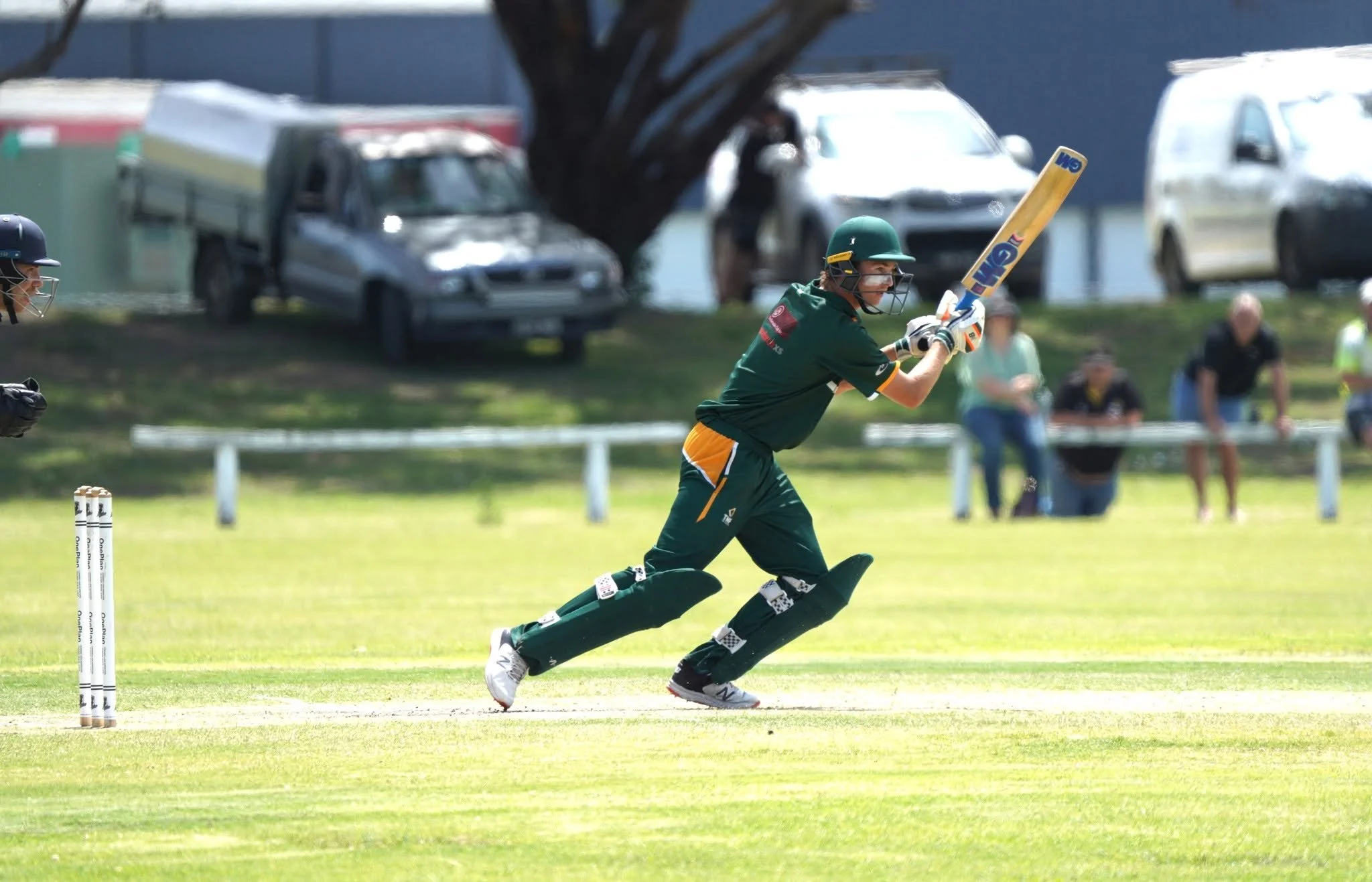 A female cricket player wearing a green and yellow uniform, helmet, and pads, running on the field while holding a cricket bat, with game spectators and parked cars in the background.