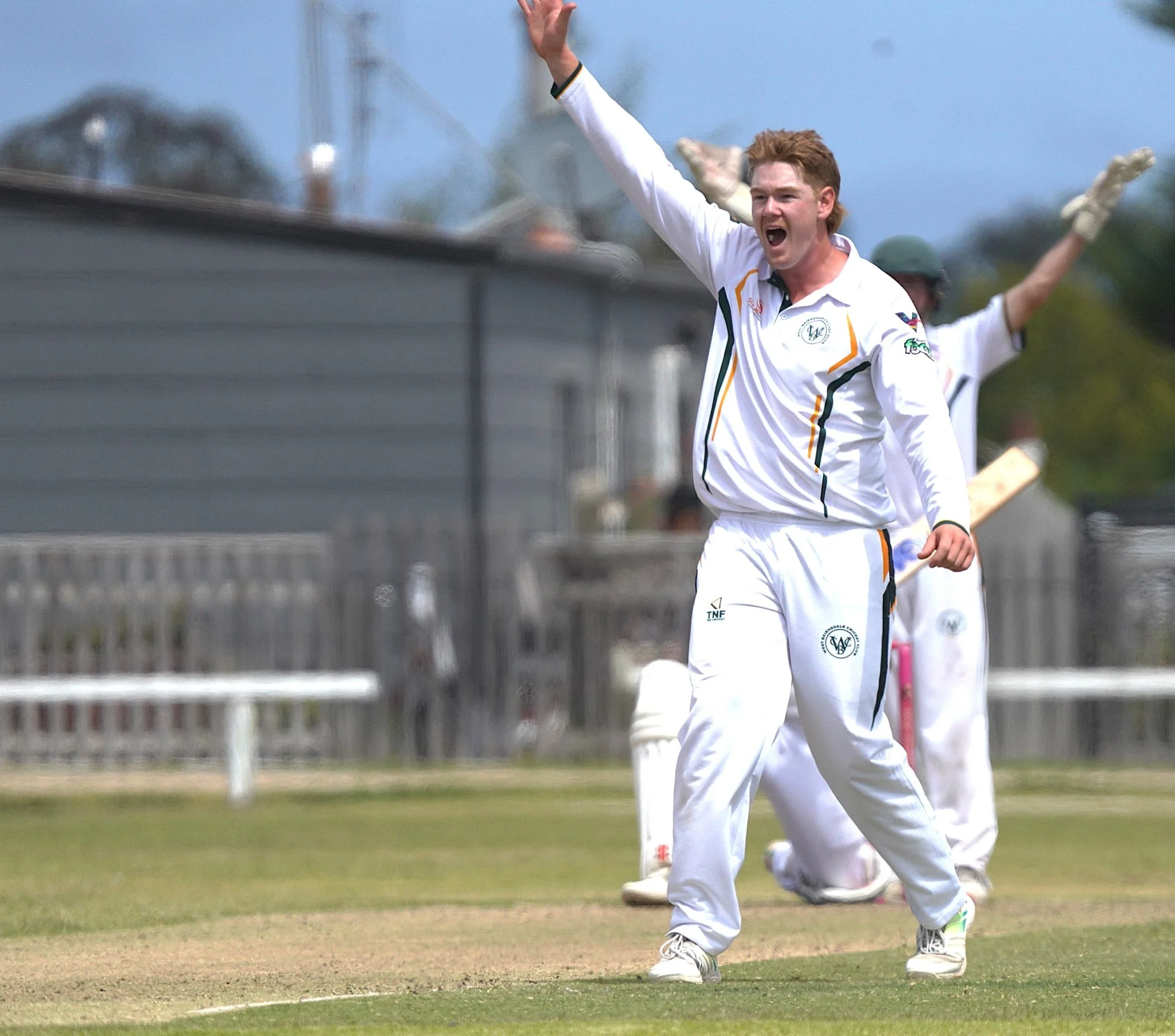 A cricket player dressed in white uniform celebrating on the cricket field with his right arm raised and mouth open, likely after taking a wicket or a successful play.