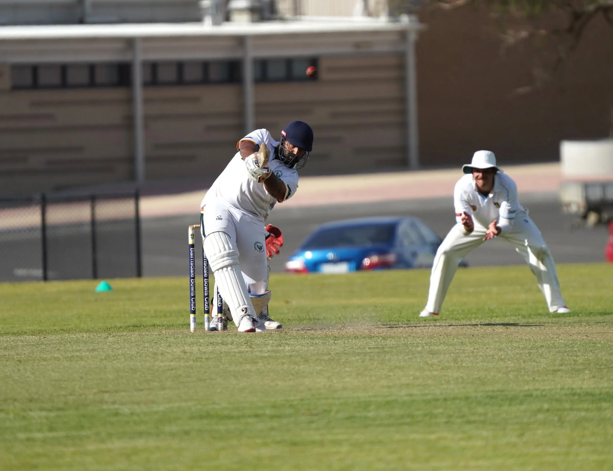 Cricket players on the field, one hitting a ball while the others are ready to catch or support, during a game on a grassy field with a building and cars in the background.