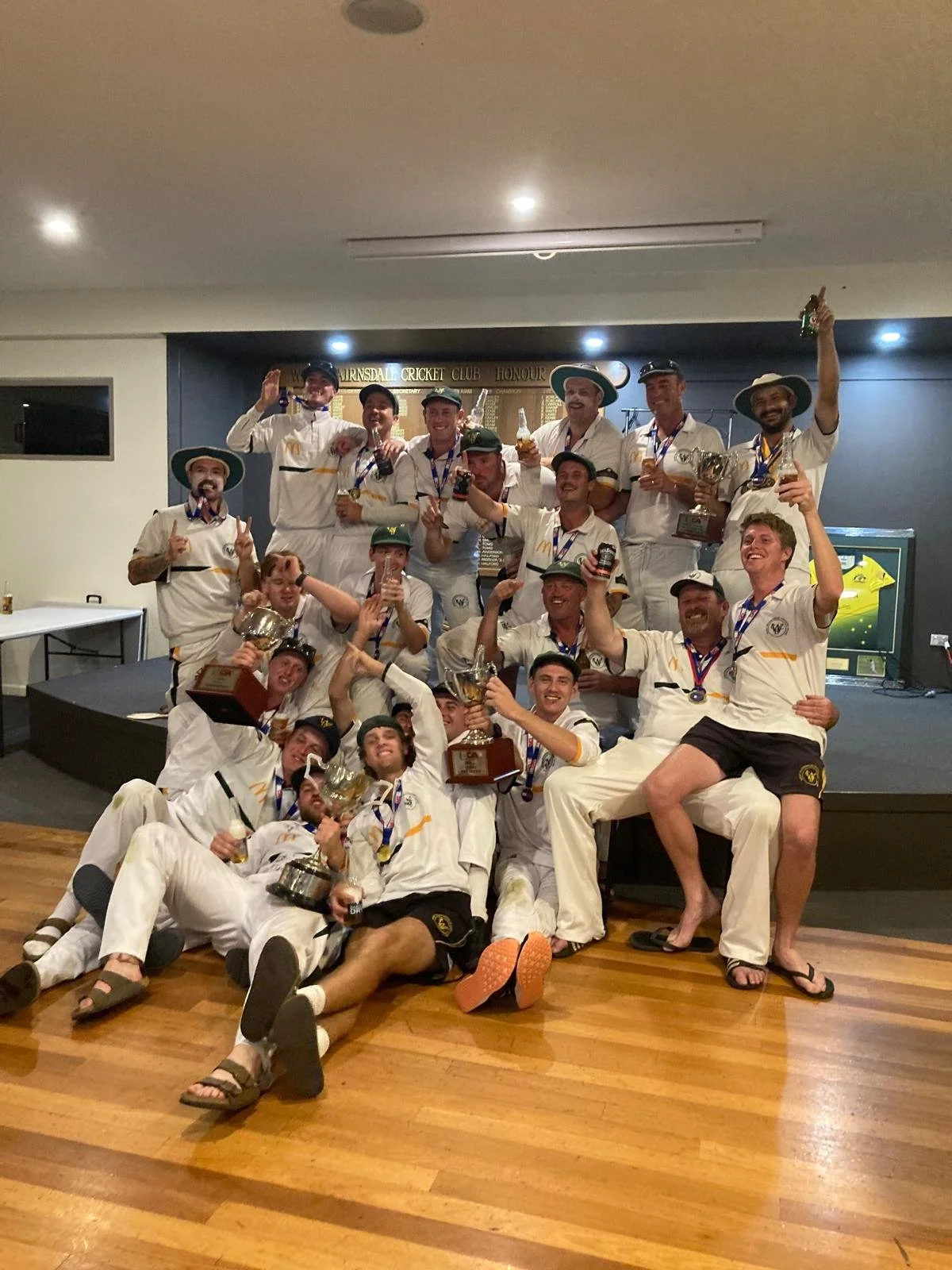 A group of cricket players celebrating their victory on a stage, holding trophies, medals, and drinks, with smiles and gestures of celebration.