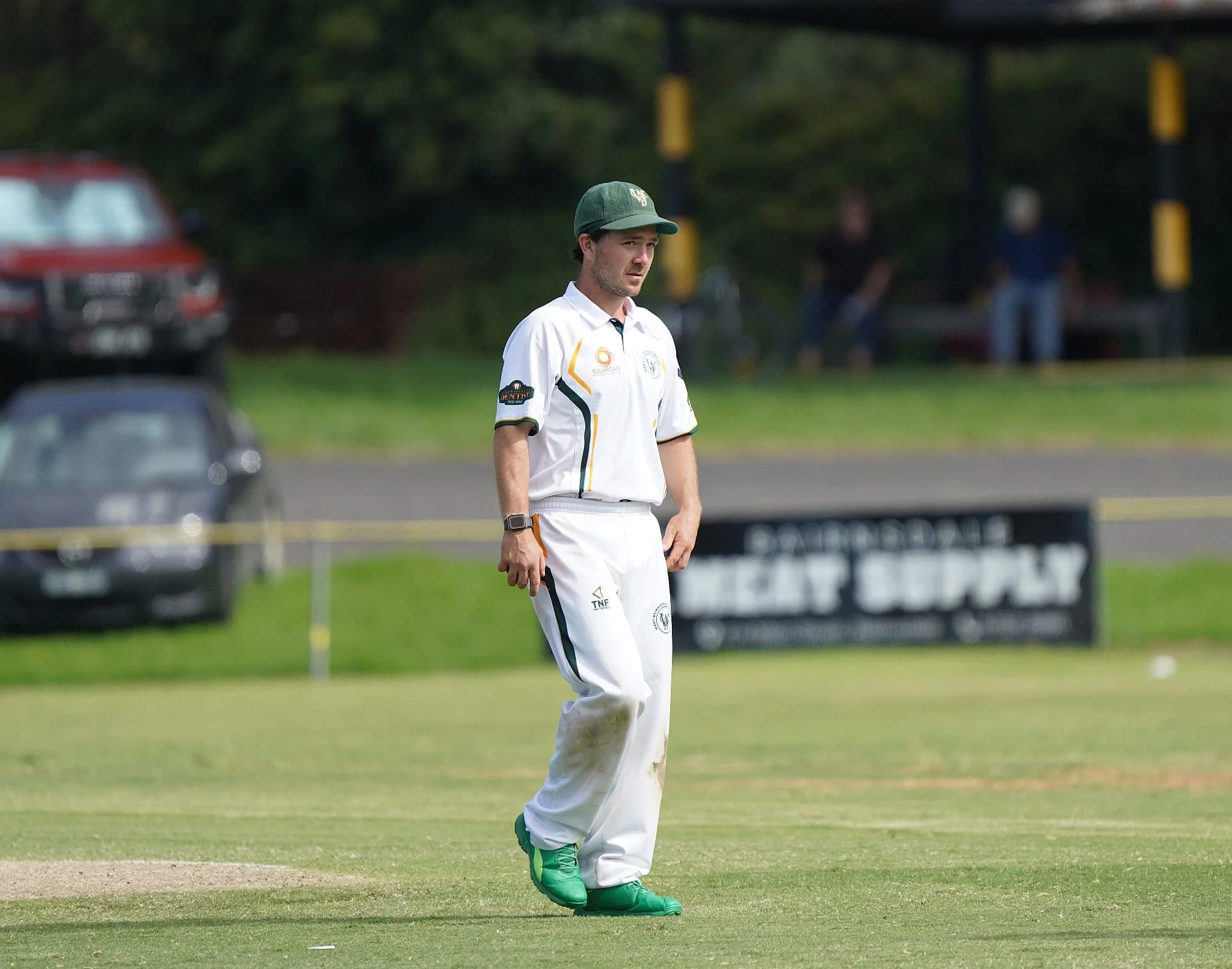 A man playing cricket on a field, wearing white cricket uniform and green sports shoes, with a dark cap and focused on the game.