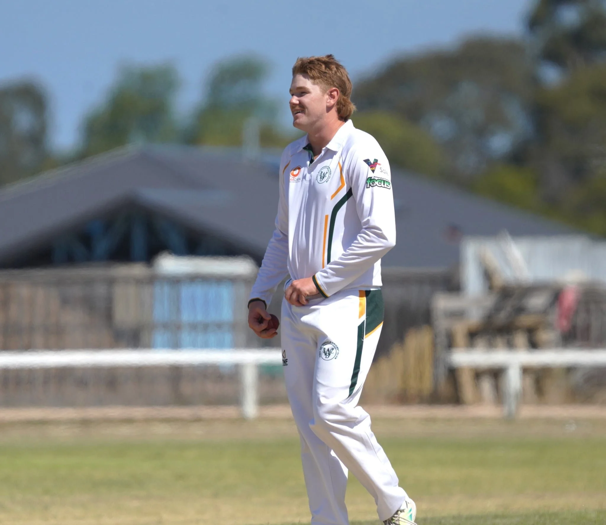 A man in a white cricket uniform standing outdoors with trees and a blue sky in the background.