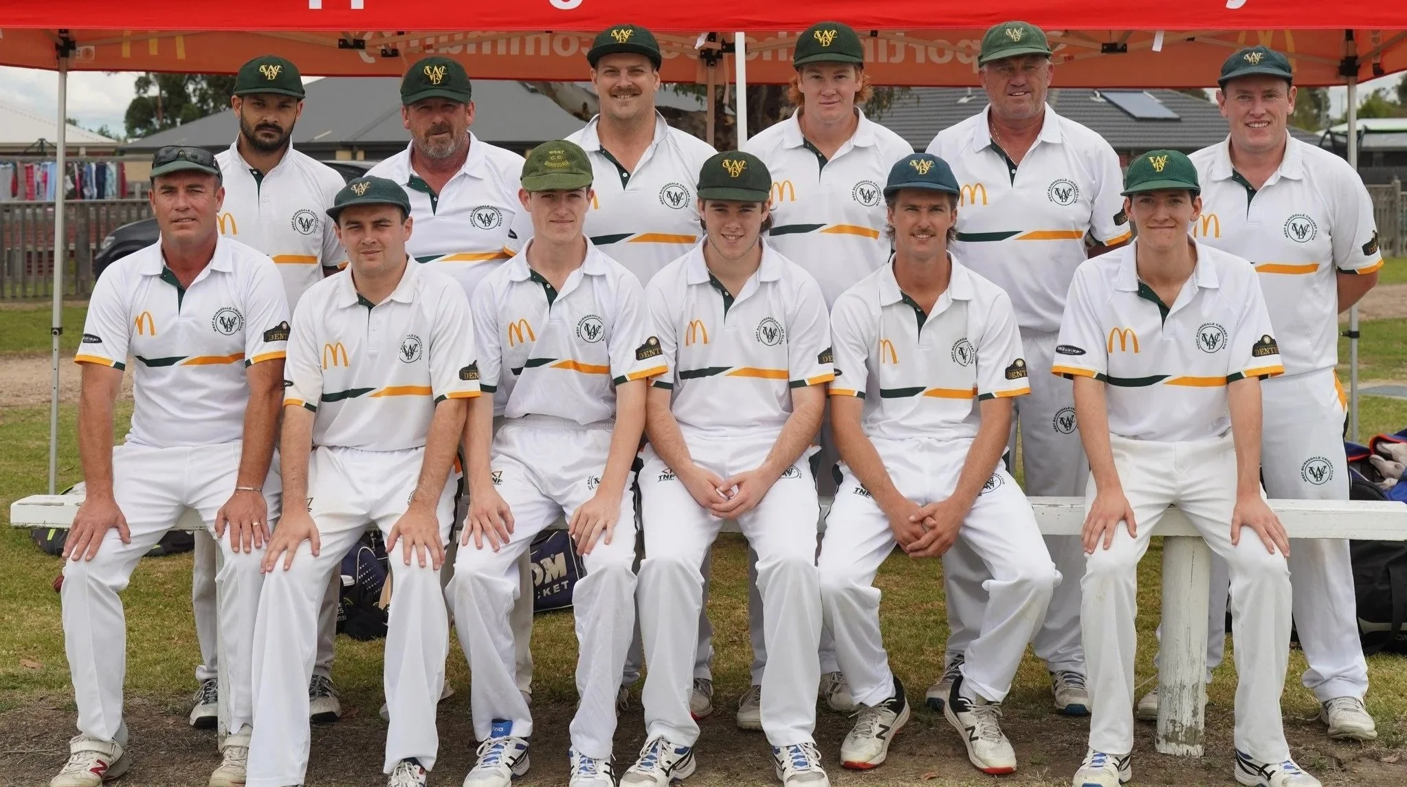 A group of cricket players in white uniforms and green caps posing together in a cricket field.