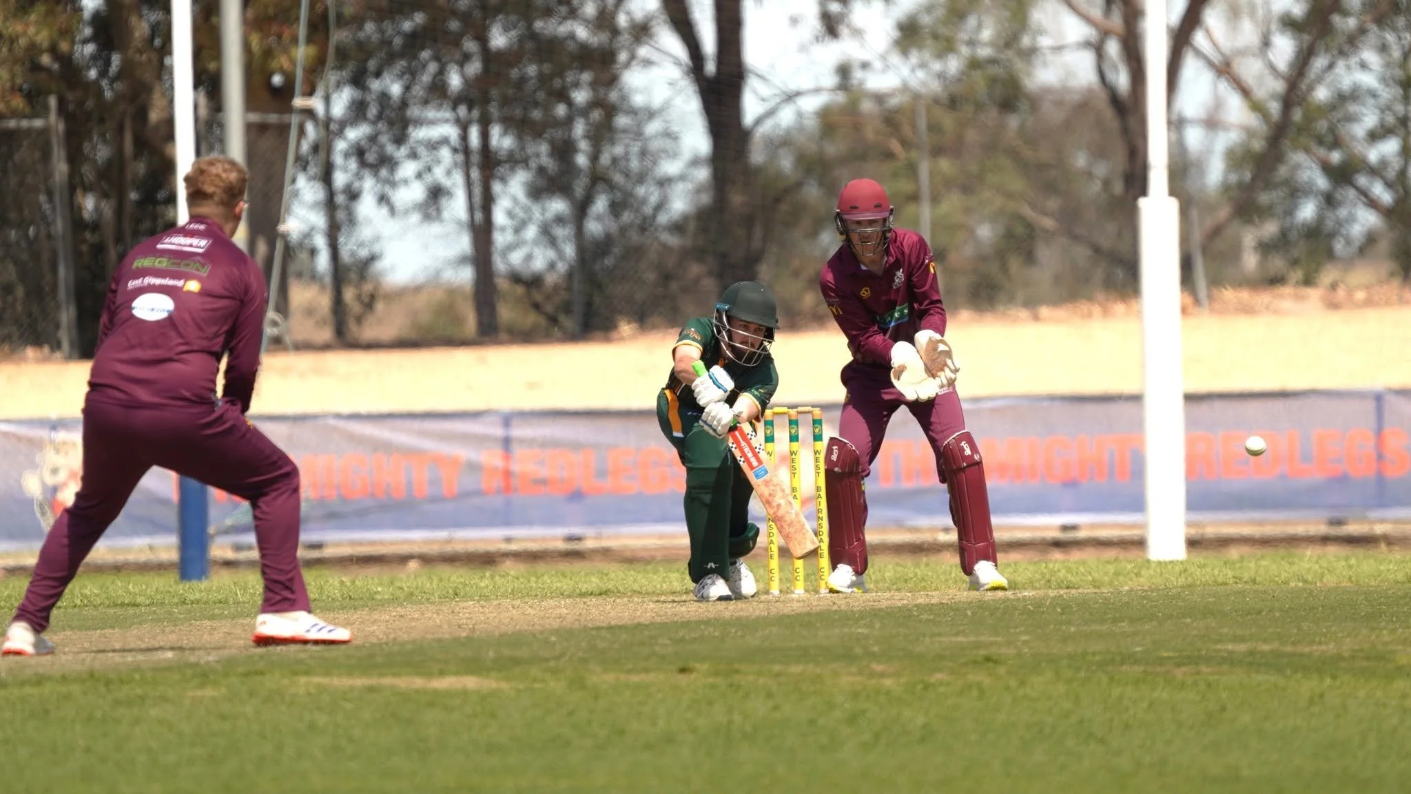 Cricket game with players on the field, one wicket keeper preparing to catch the ball, and an opposing player approaching the wicket, on a cricket pitch with dry grass and trees in the background.