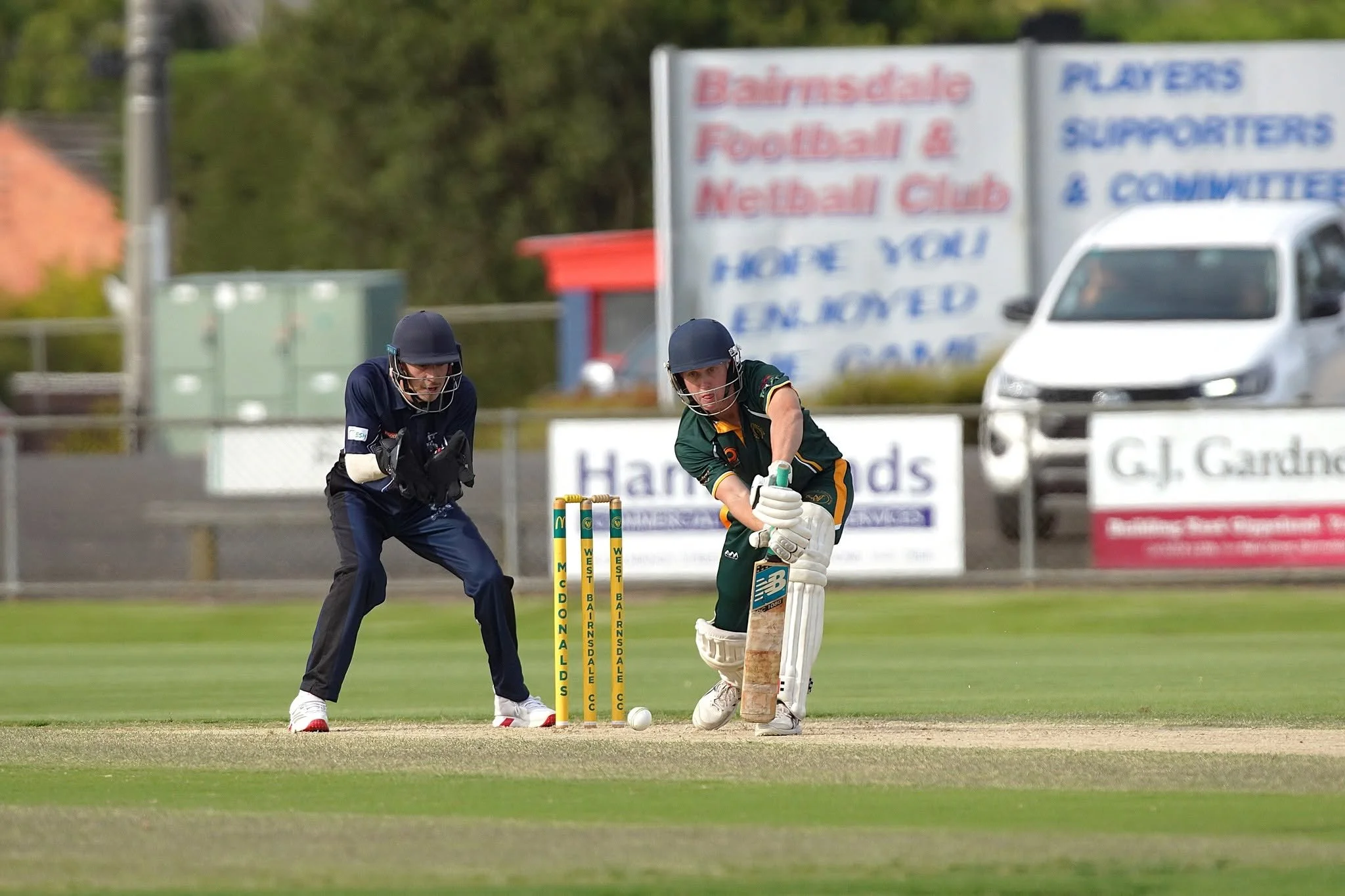 Cricket match with two players near wickets on a grassy field, with advertising and trees in the background.