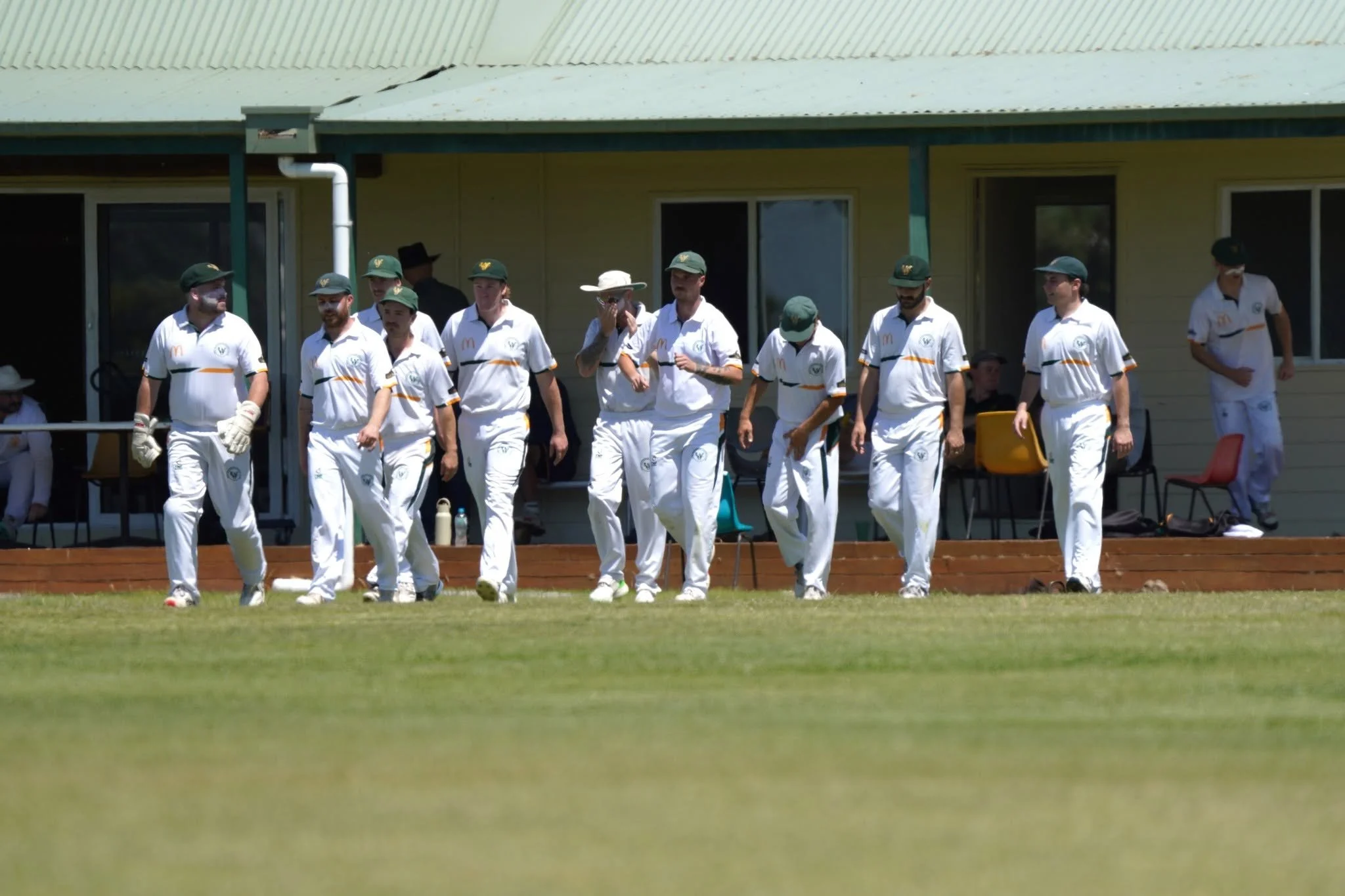 A group of cricket players in uniform walking on a grassy field, with a few seated and standing spectators in the background.