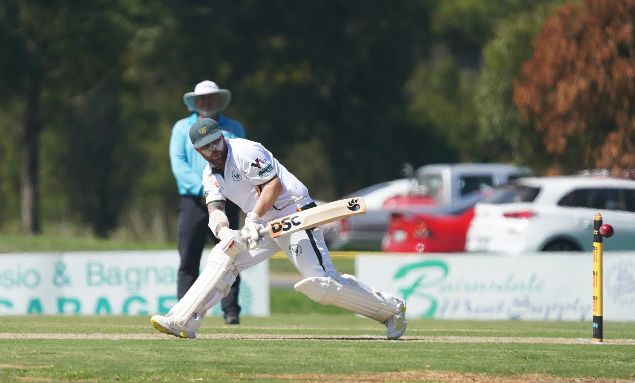 A cricket player in white uniform diving forward while trying to catch a ball during a game, with an umpire and parked cars in the background.