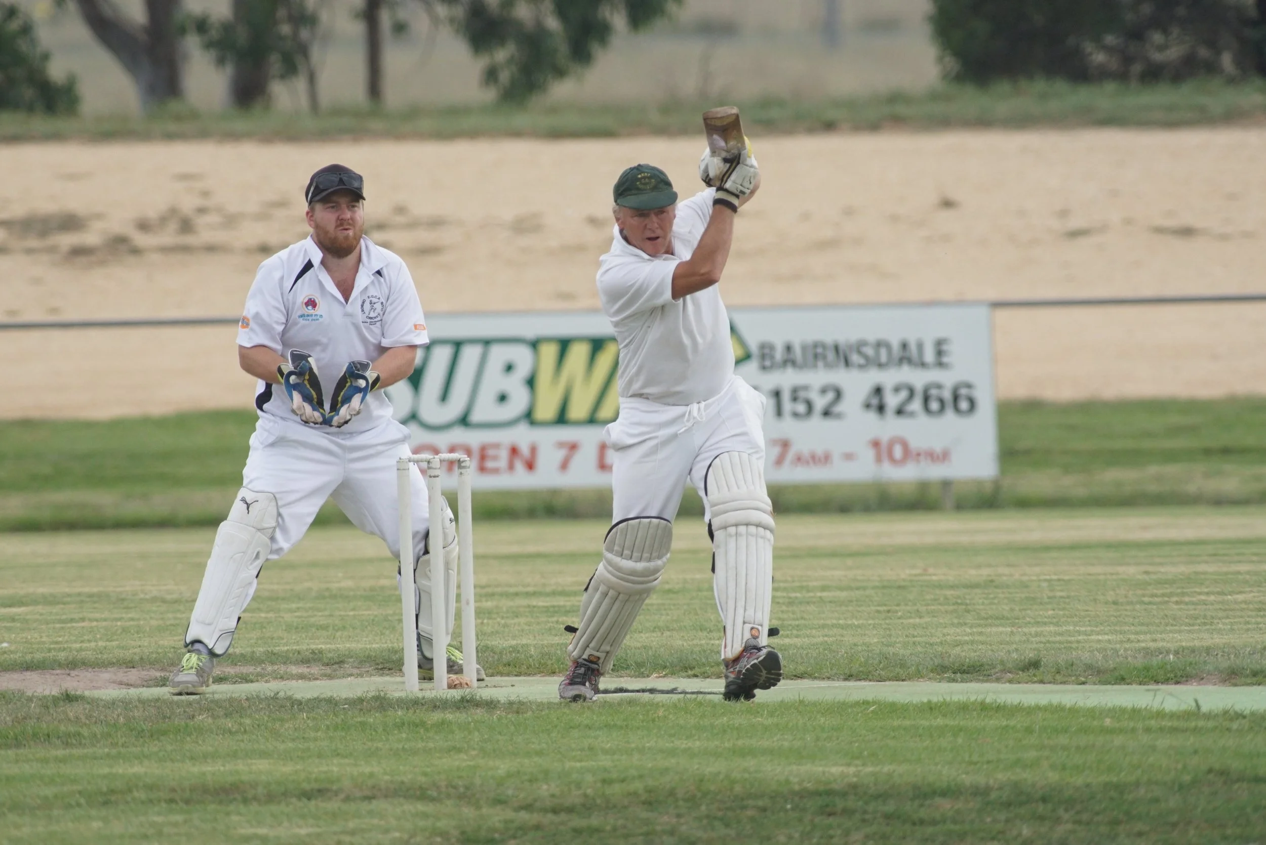 Two cricket players on the field, one batting and the other wicketkeeping, during a match on a grassy cricket ground, with trees and advertising signs in the background.