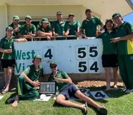 Group of ten people celebrating a lawn bowls game at a bowling club, with a scoreboard showing the West team winning 4-0 against the Vics team, and two players sitting on the grass in front with a trophy and a glass of drink.