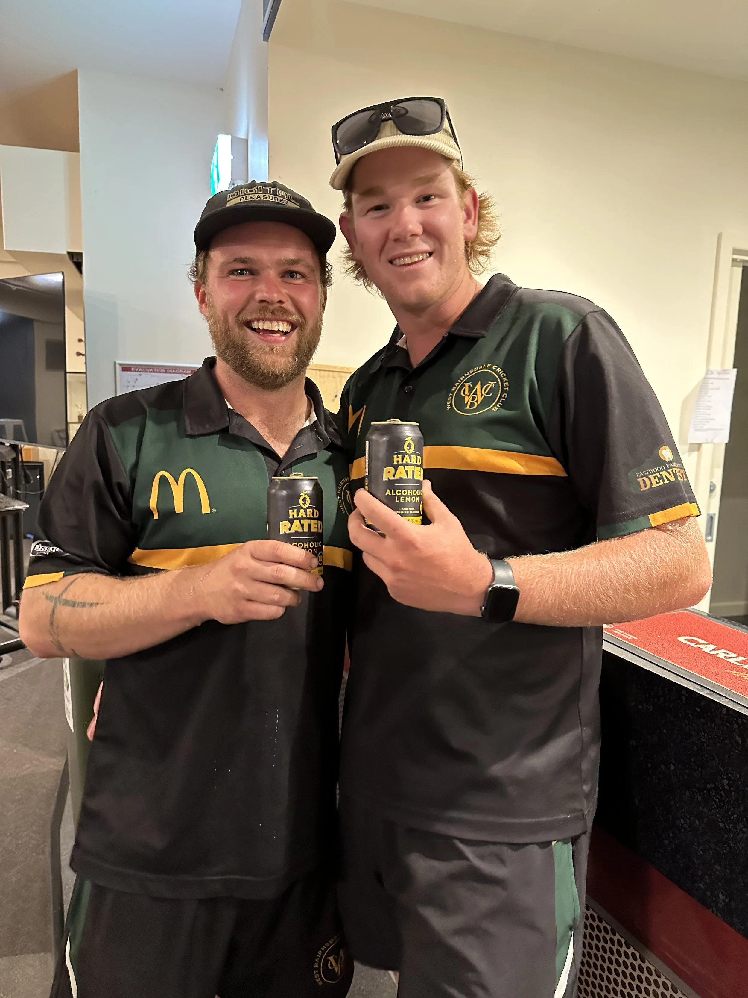Two young men smiling, holding cans of alcoholic lemon beverage, standing together indoors. Both are wearing black and green sports jerseys with Nike and other logos. One is wearing a baseball cap, sunglasses on his head, and a watch. The other has a