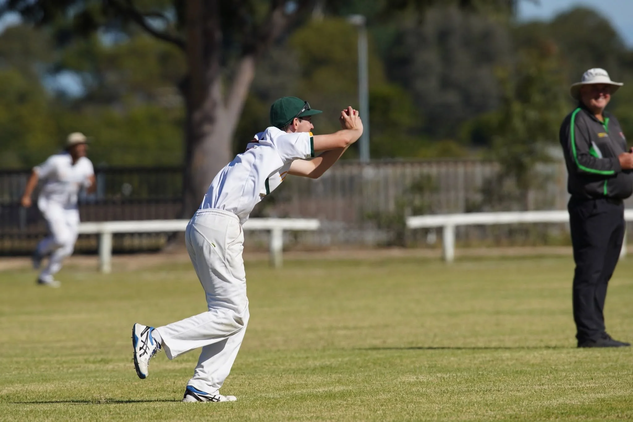 Young boy in white cricket uniform and green cap running on a field during a cricket match.