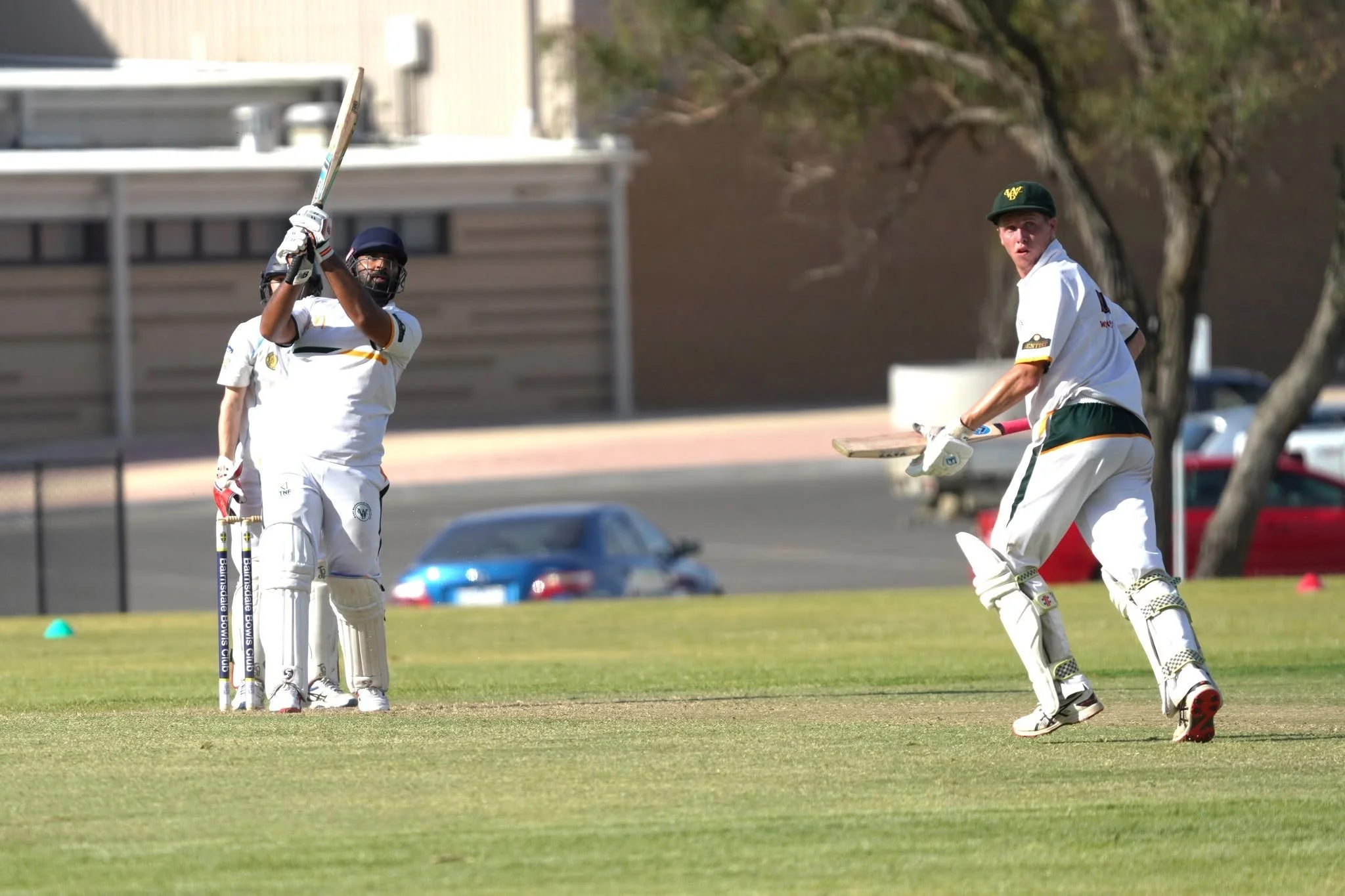 Cricket players in white uniforms playing on a grassy field, with trees and parked cars in the background.