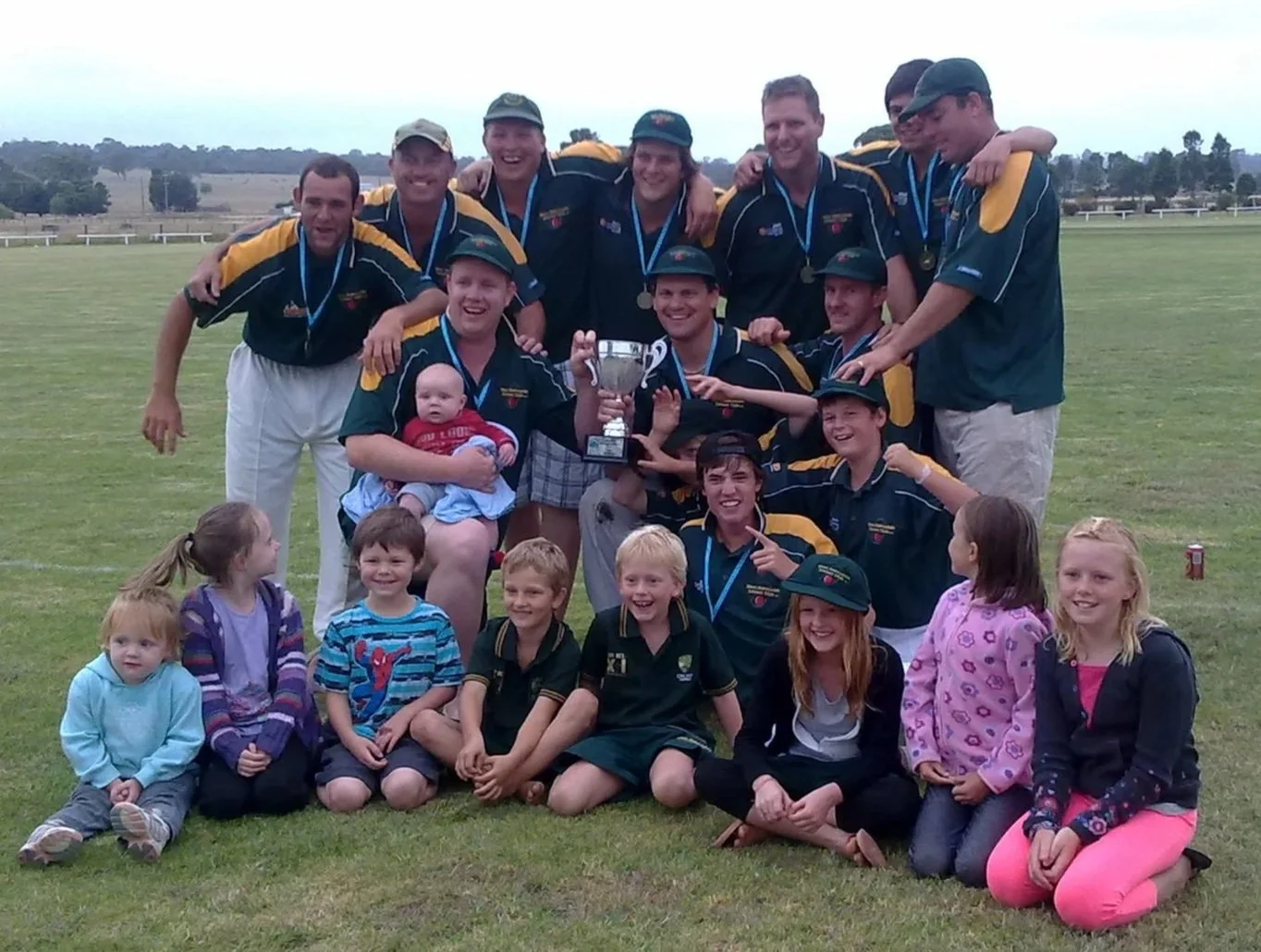 A group of children and adults celebrating a sports victory on a grassy field, with some holding a trophy and medals, and kids sitting in front smiling.