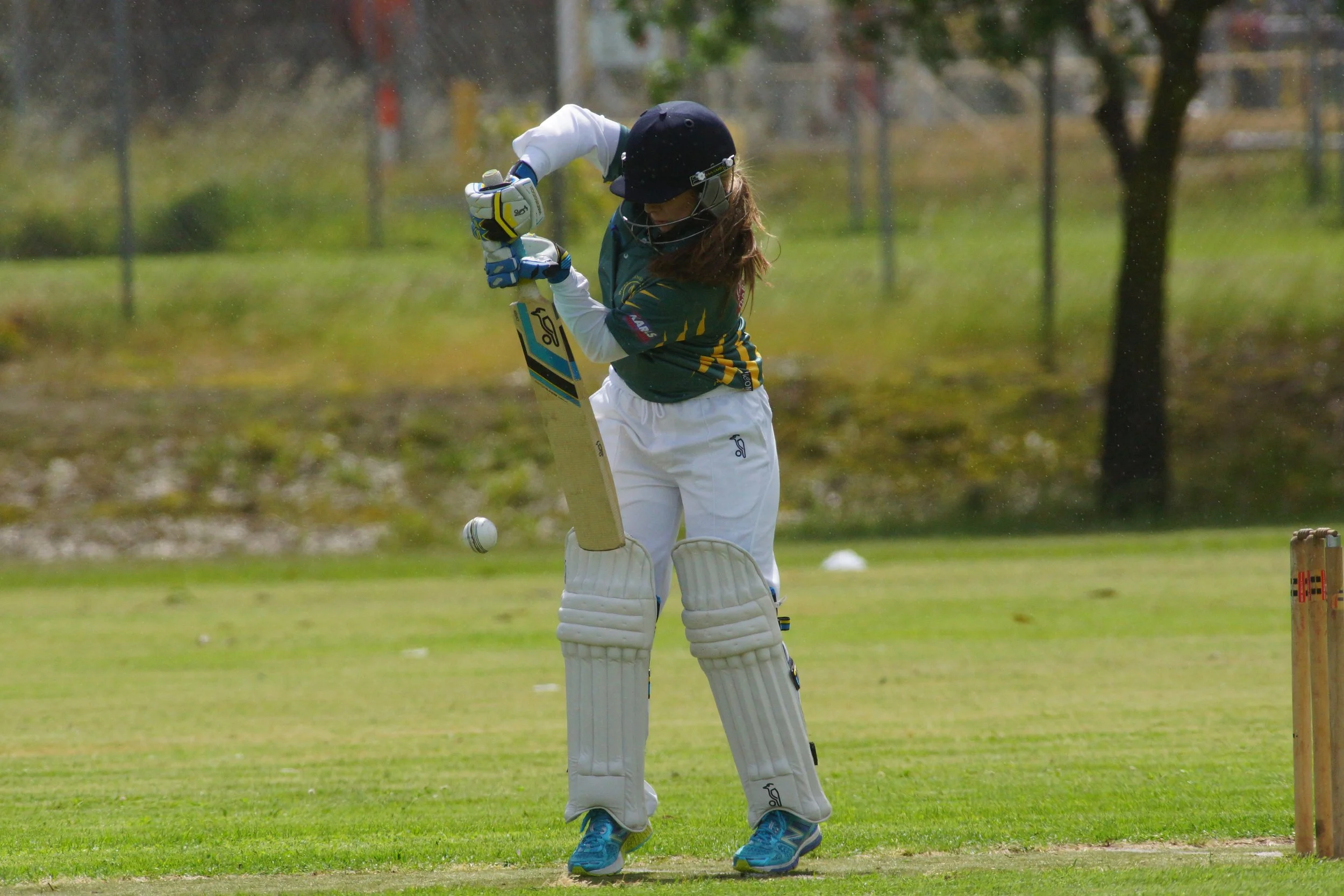 A female cricket player in full gear, including pads, gloves, and helmet, is swinging a cricket bat at a ball on a cricket field during daytime.