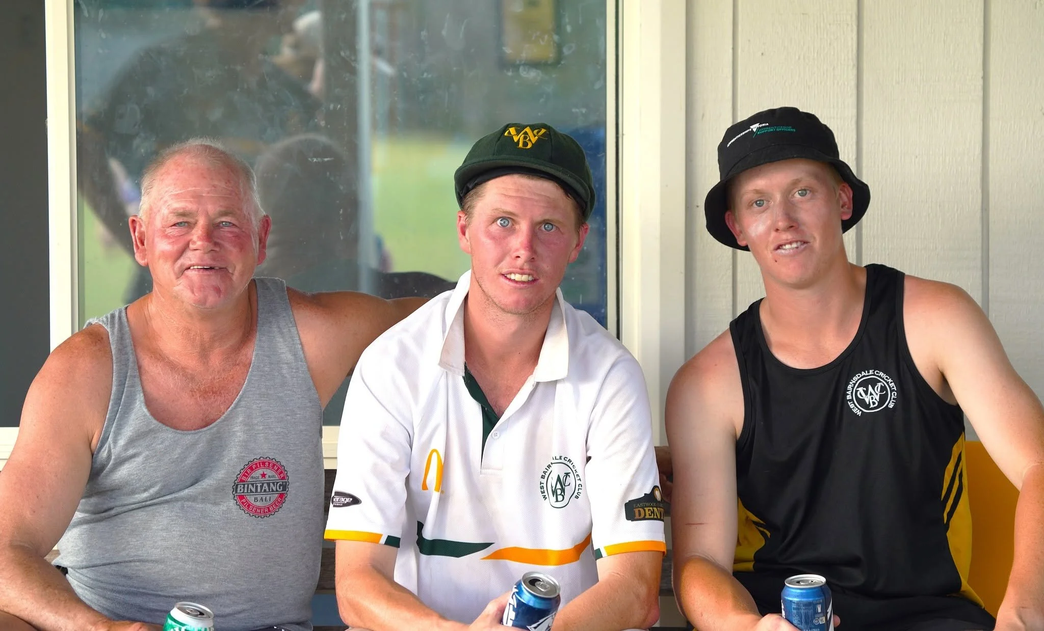 Three men sitting side by side, smiling at the camera. The man on the left is older, wearing a gray tank top. The man in the middle is younger, wearing a white sports shirt with green and orange accents and a black cap with yellow writing, holding a 