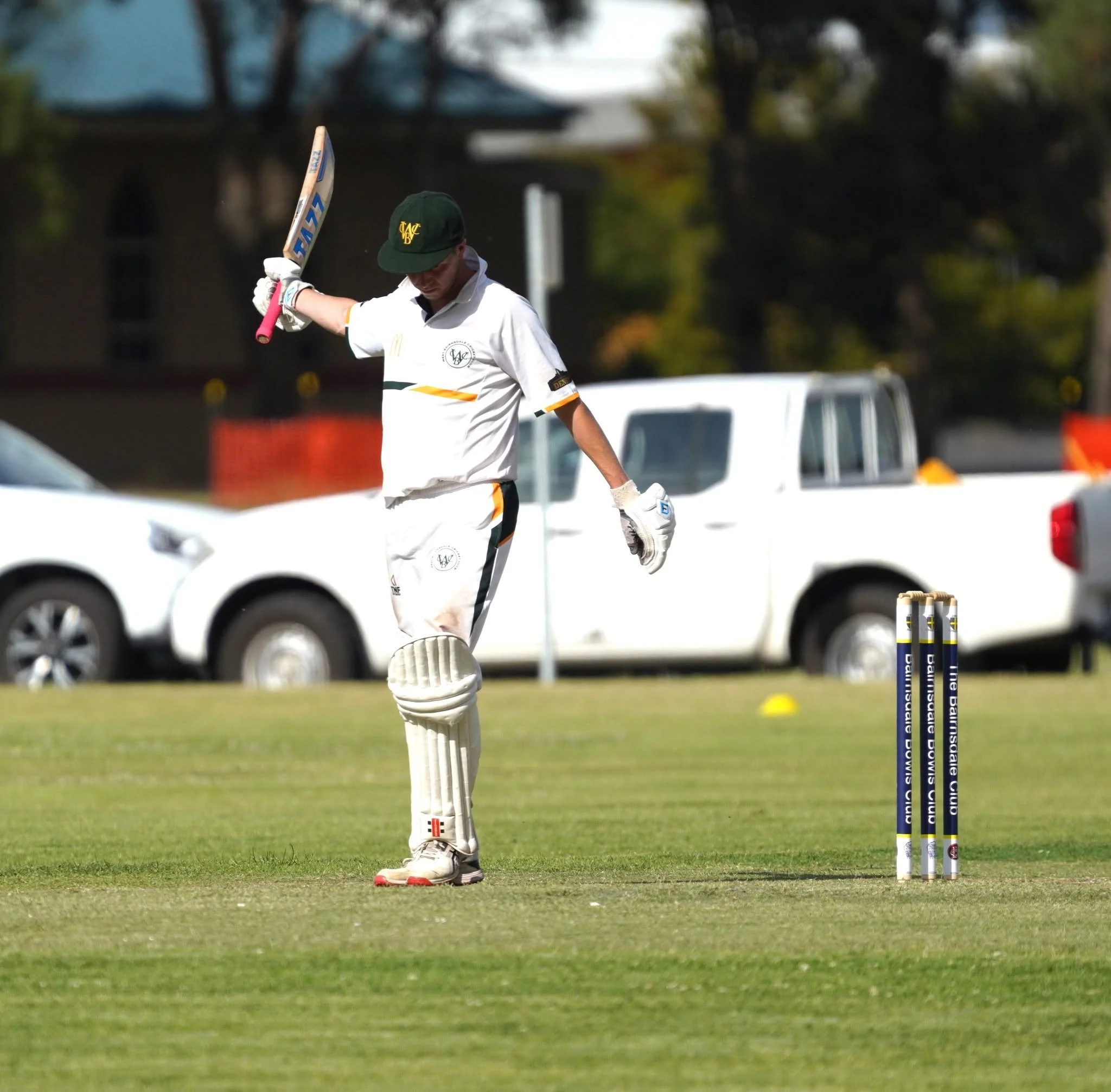 A cricket player in white uniform and black cap, holding a cricket bat and celebrating on a green field, with stumps in front and a blurred background of cars and trees.