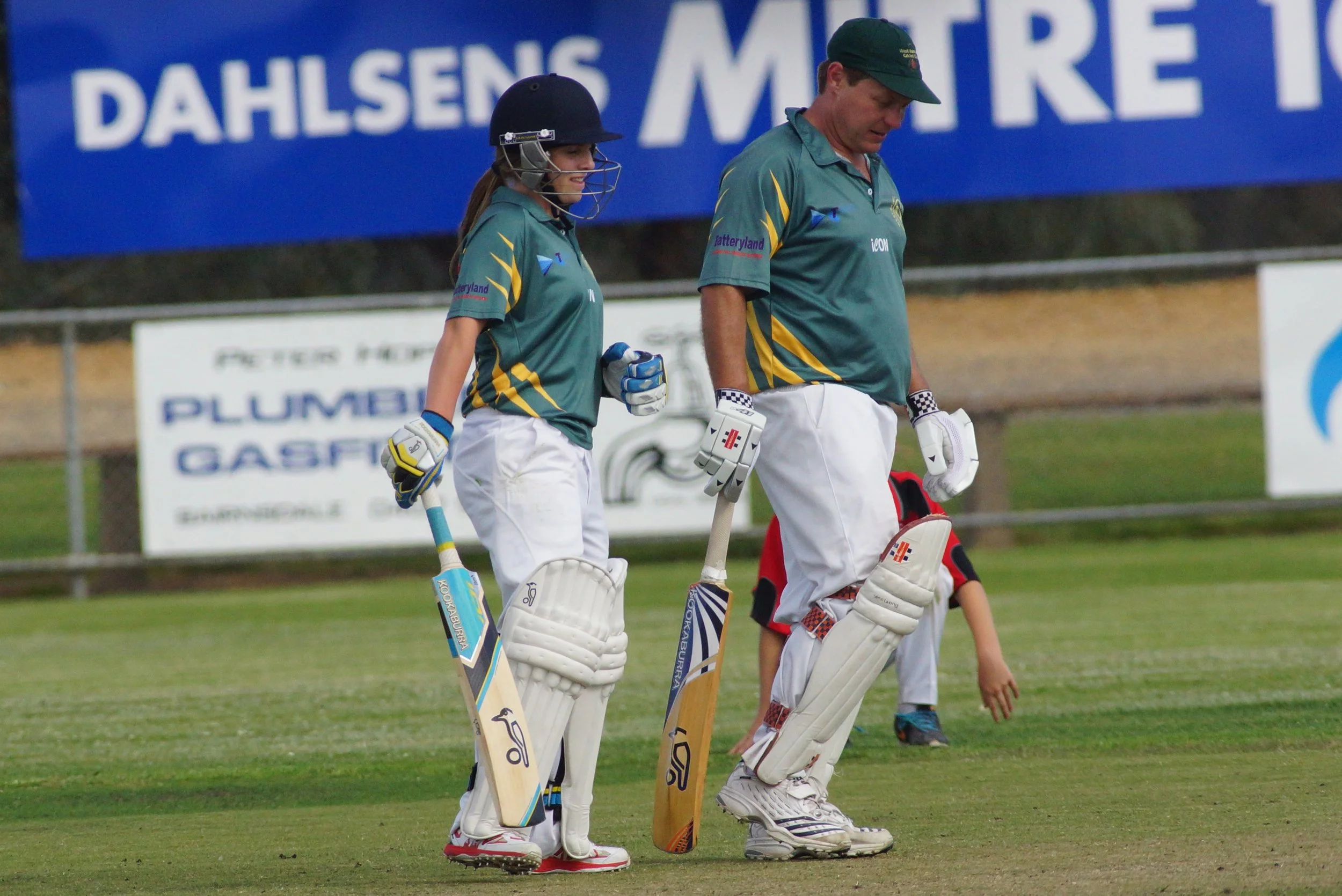 Two cricket players and a young boy walking on the field, with a blue banner in the background.