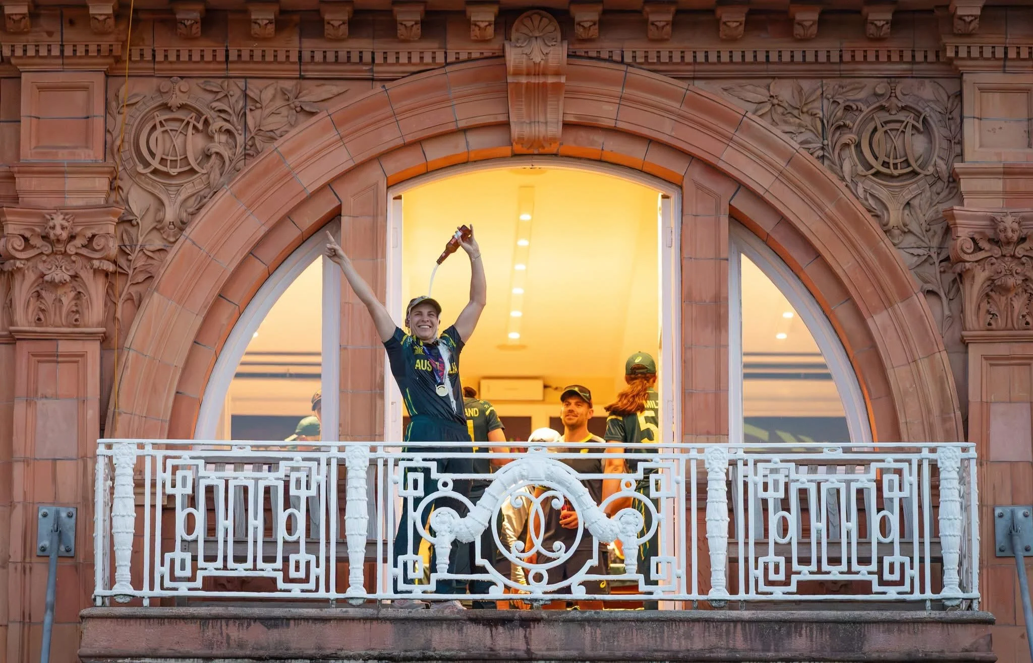 A group of Australian rugby players celebrating on a balcony after a match, with one player holding a trophy, wearing medals, and smiling.