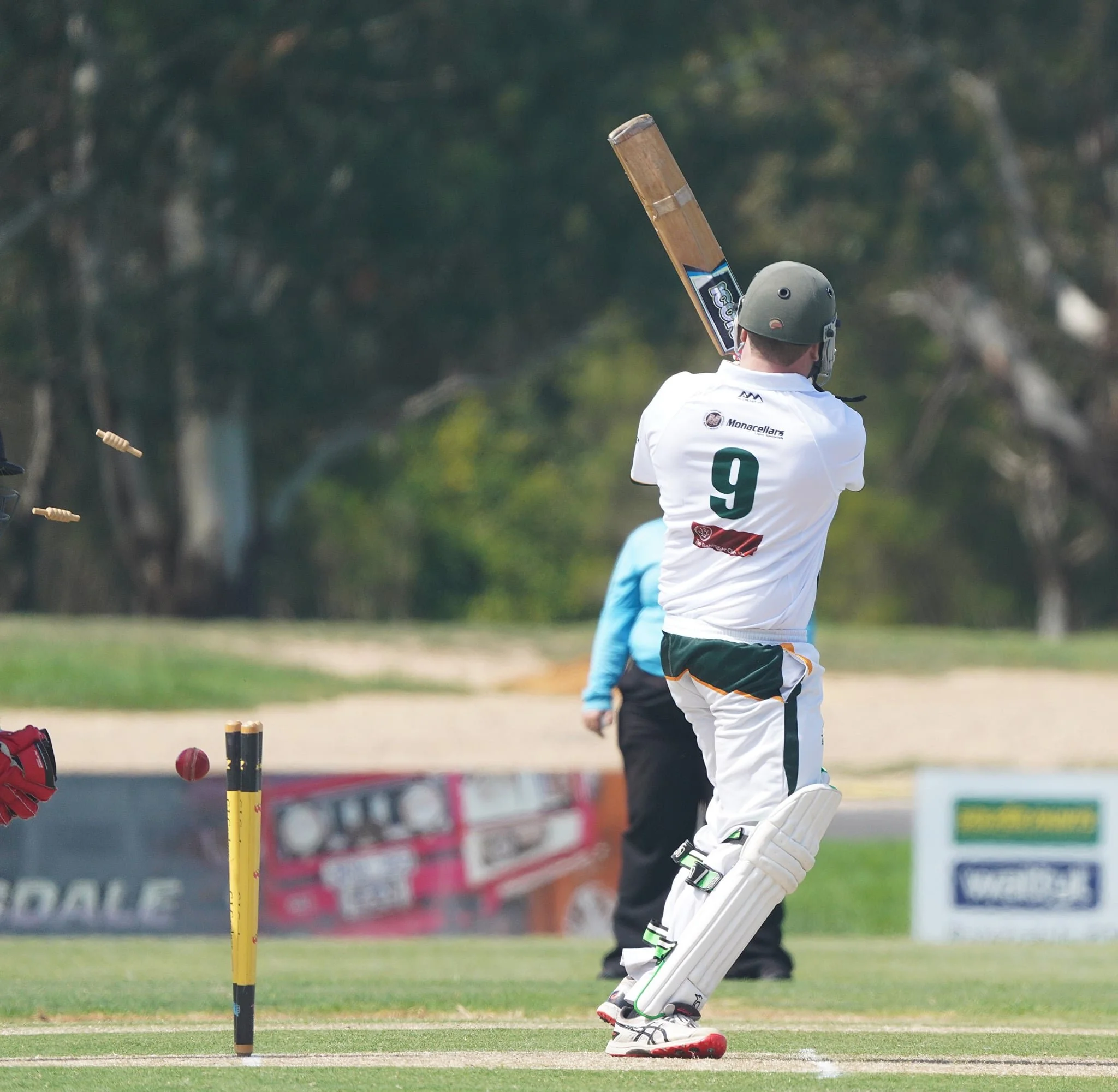 Cricket player in white uniform and gray helmet hitting the ball with a cricket bat during a game on a grassy field with trees in the background.