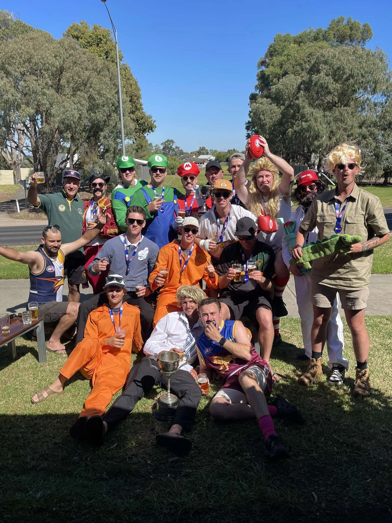 Group of people dressed in costume celebrating outdoors, some holding drinks, others with medals and trophies, with trees and a clear blue sky in the background.