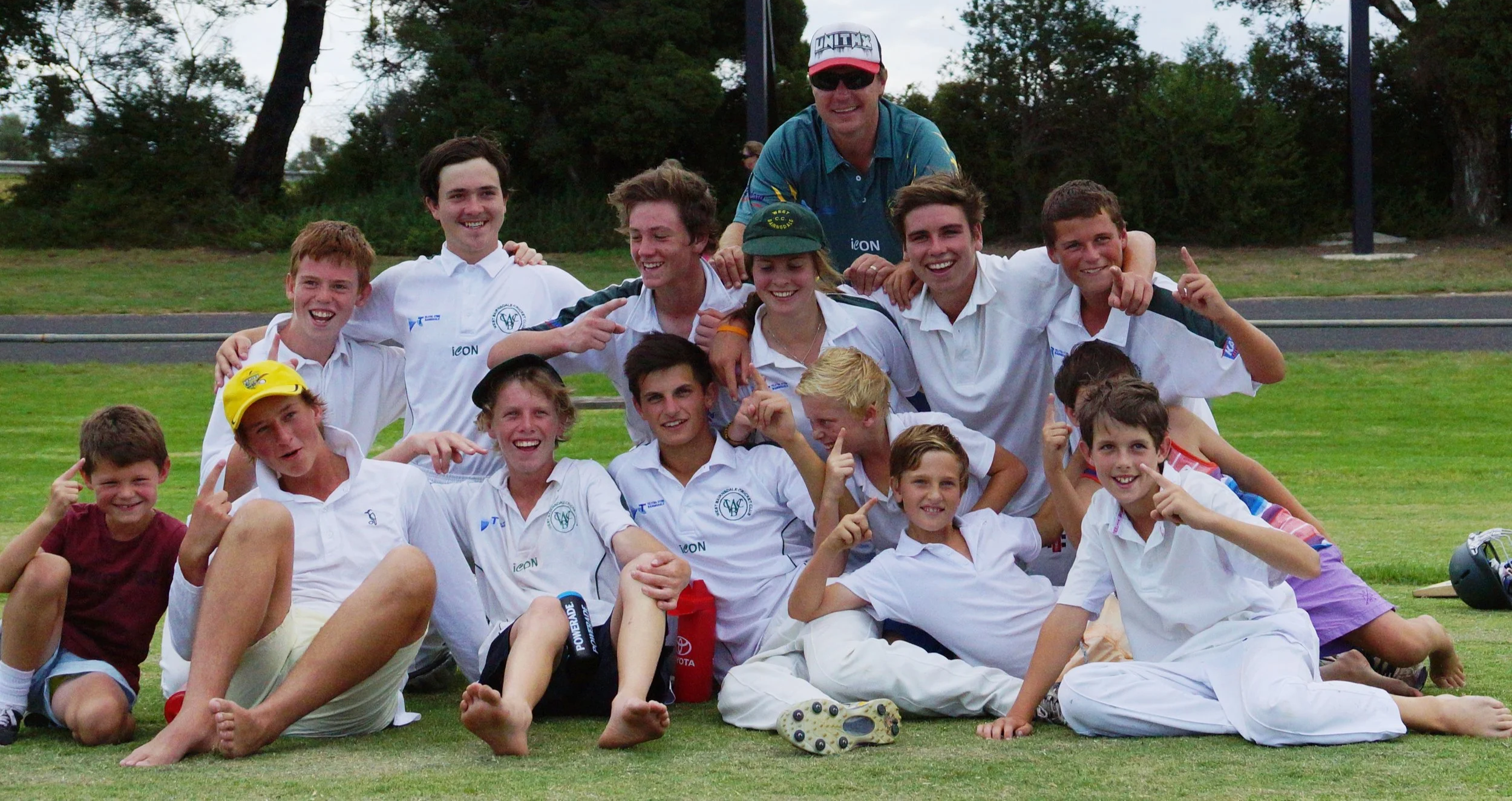 A group of young boys and a few adults posing outdoors on a grassy field, all smiling and making playful gestures.