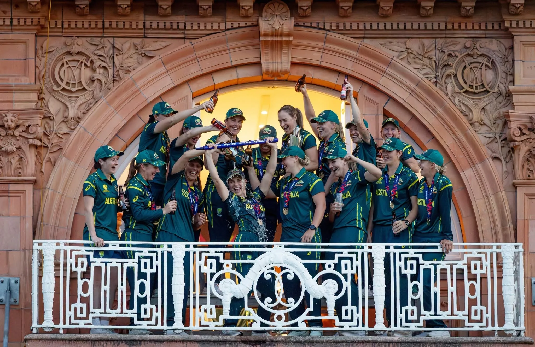 Group of female Australian athletes celebrating on a balcony after winning a competition, holding bottles and medals, wearing green and yellow sportswear and caps, with a historic building with ornate architectural details in the background.