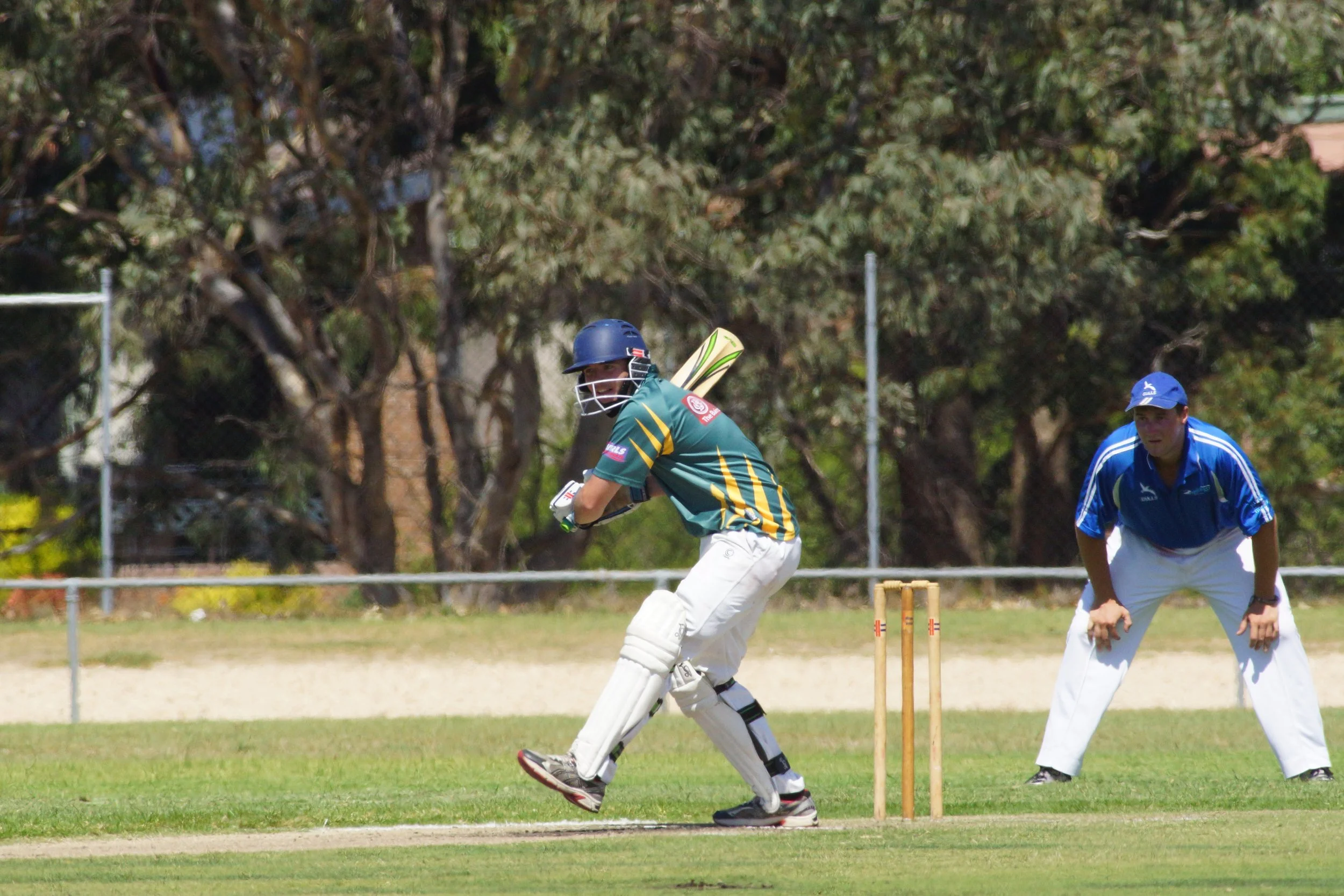 Two men playing cricket outdoors on a grassy field, one of whom is batting and wearing a helmet, and the other is fielding nearby, with trees and fencing in the background.