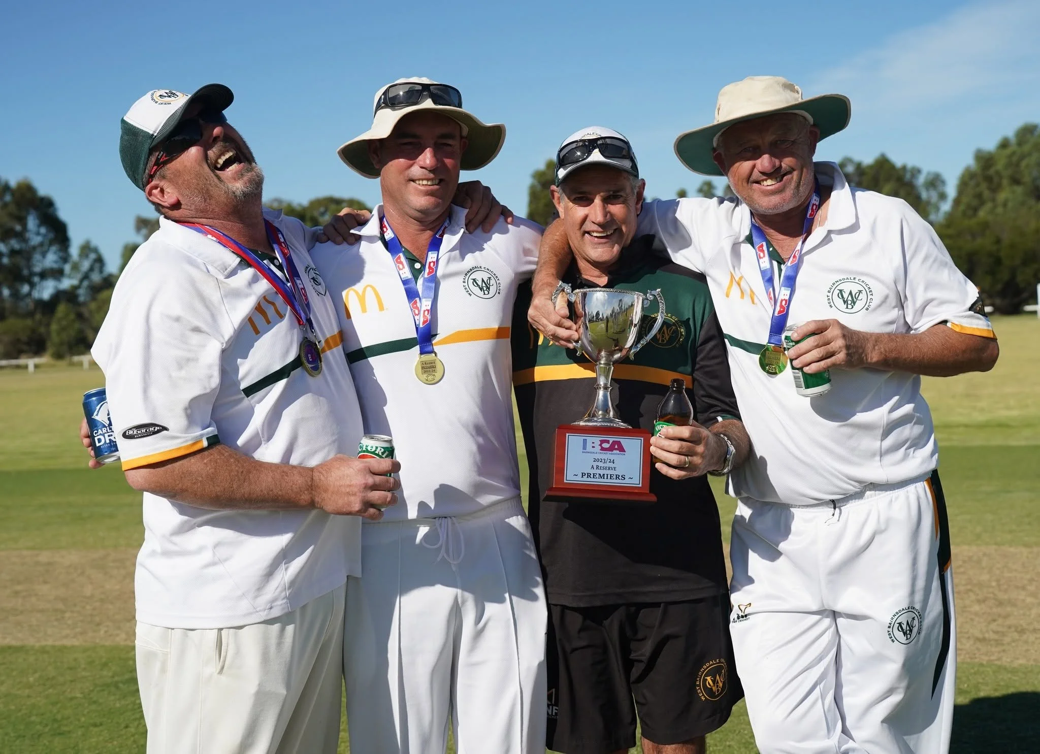 Four men celebrating on a cricket field, holding a trophy and wearing medals, with a clear blue sky and trees in the background.