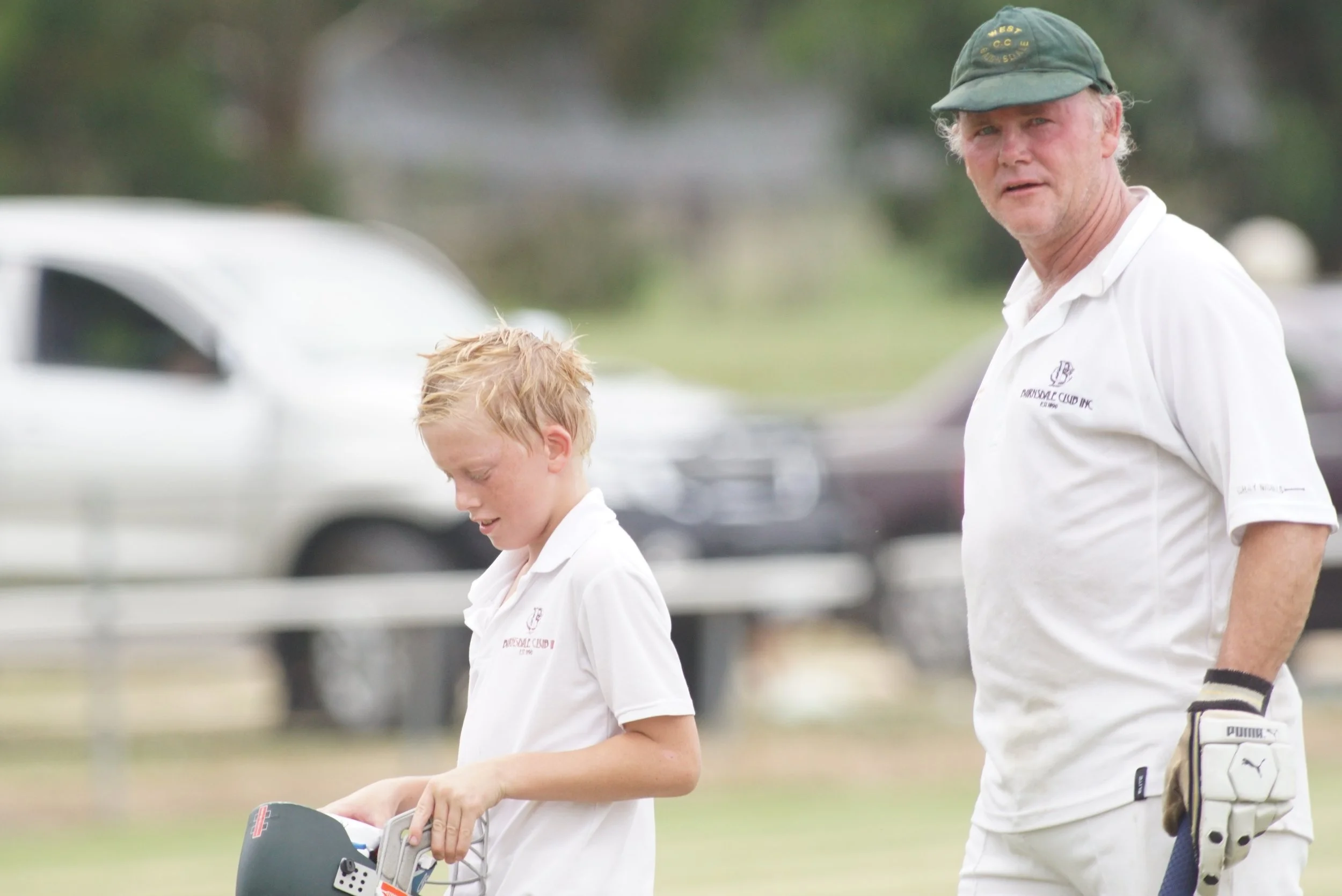 A man and a young boy in white shirts walking outdoors near parked cars, with the man wearing a cap and the boy holding a helmet.