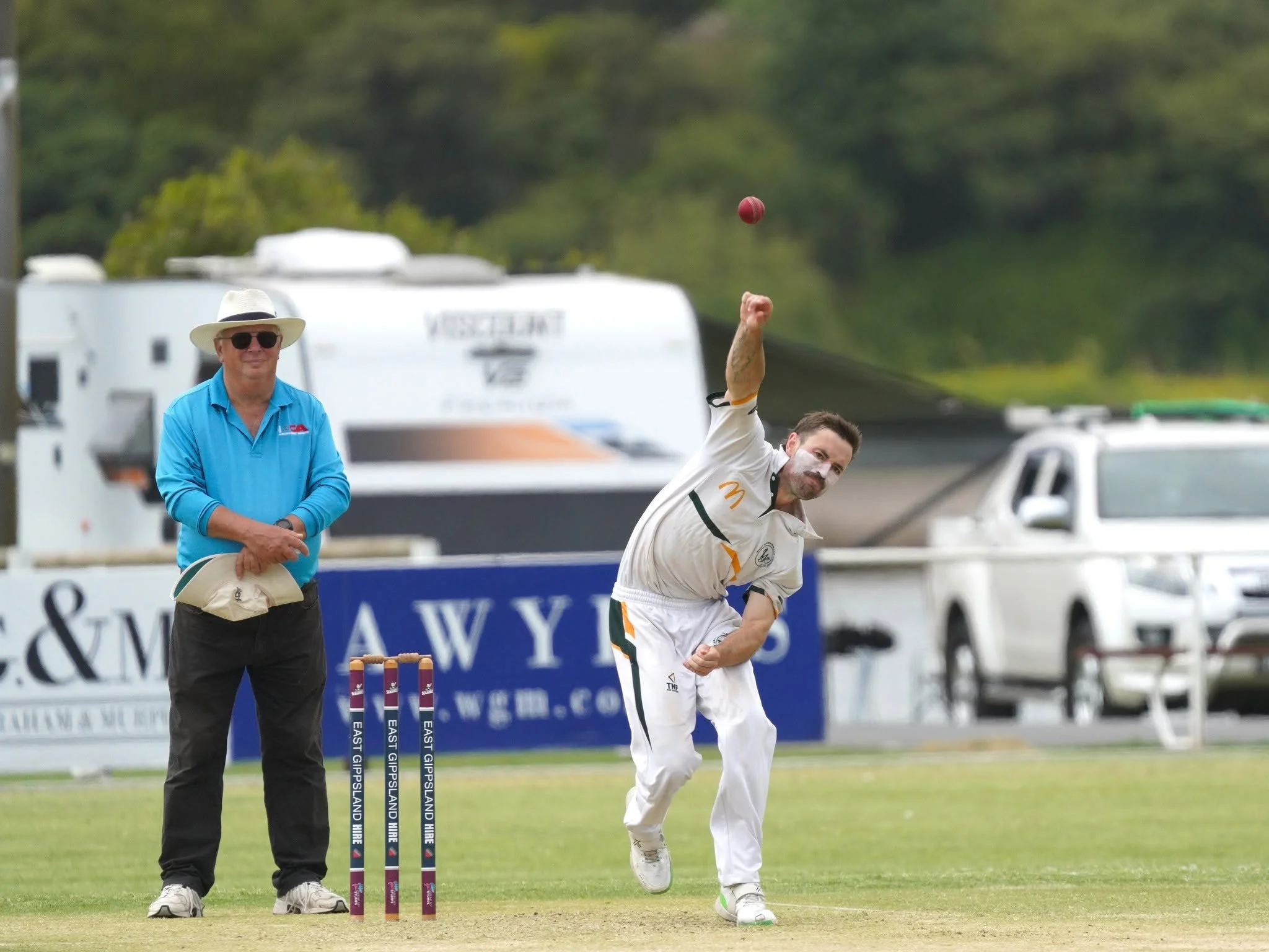 A cricket player in white uniform and green shoes is celebrating after scoring runs, lifting his arm, while an umpire in a blue shirt, black pants, white hat, and sunglasses looks on. There are advertisement banners and a cricket stumps with the bran