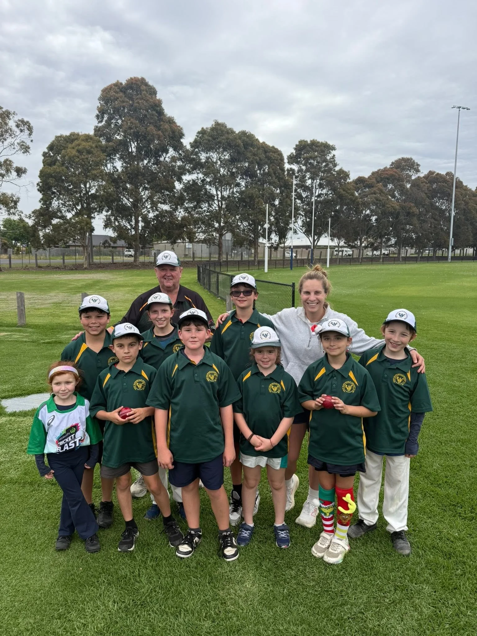 A group of children and two adults posing on a sports field. The children are wearing green sports jerseys and hats, while the adults are dressed casually. The group appears to be part of a cricket team, with some children holding cricket balls. The 