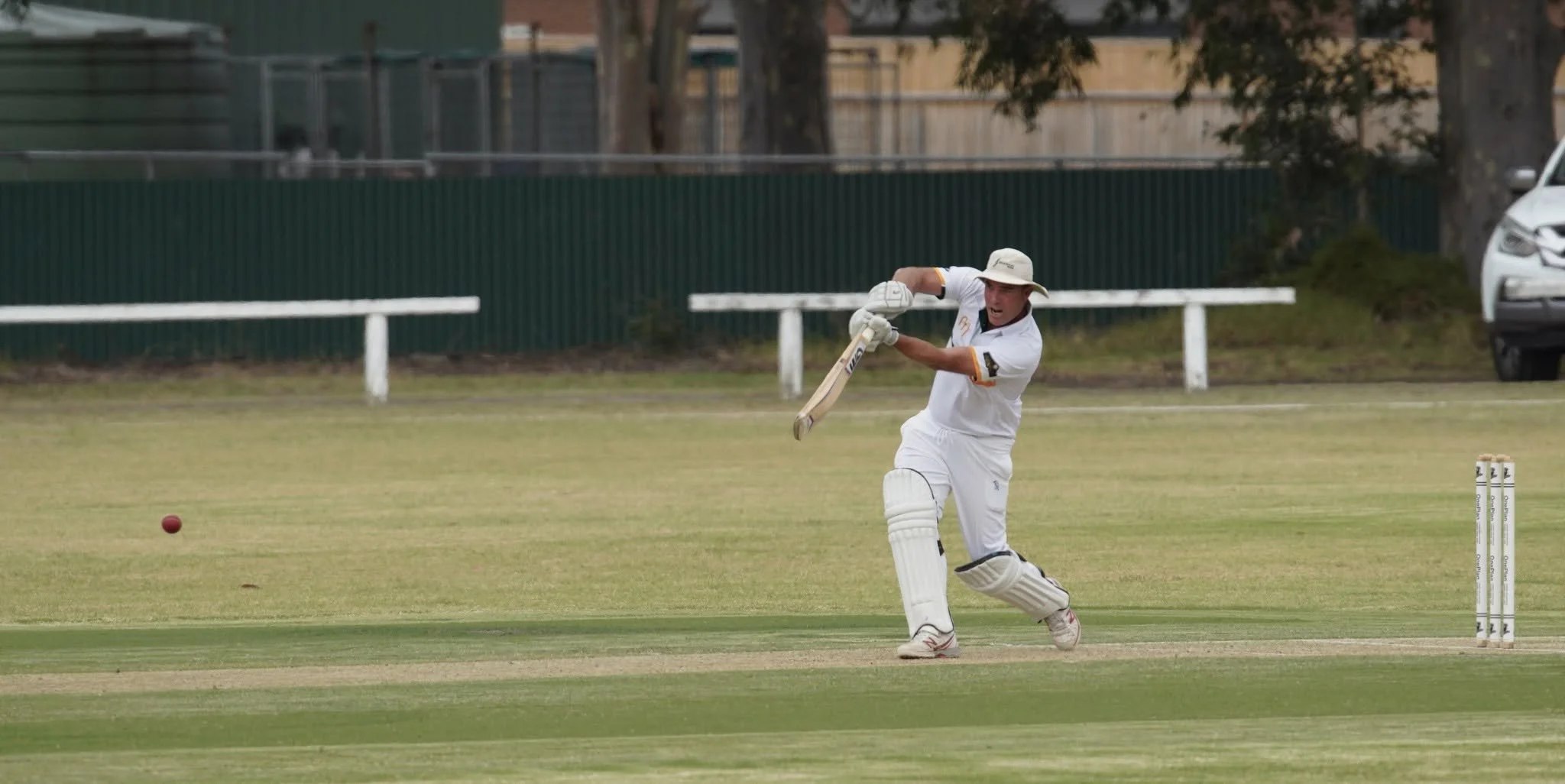 A cricket player in white uniform and a wide-brimmed hat is hitting a cricket ball with a bat on a grass field, with wickets nearby and a car and trees in the background.
