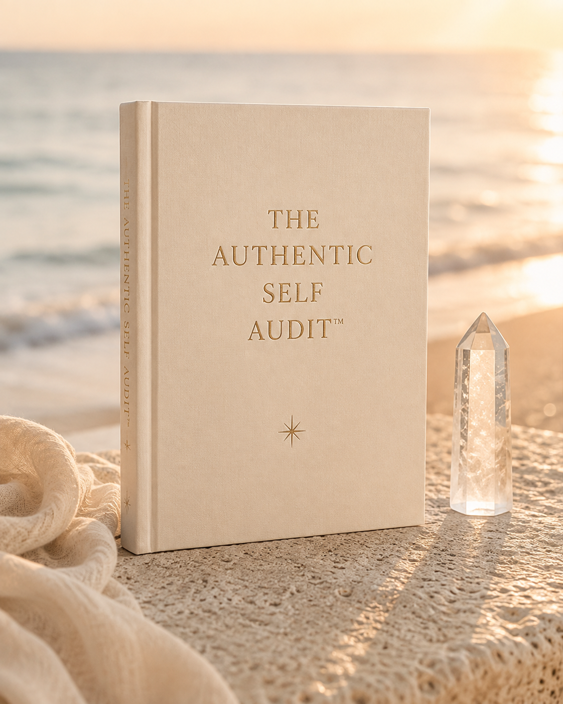 A beige book titled 'The Authentic Self Audit' standing on a textured surface near the ocean at sunset, with a clear quartz crystal and a piece of fabric nearby.