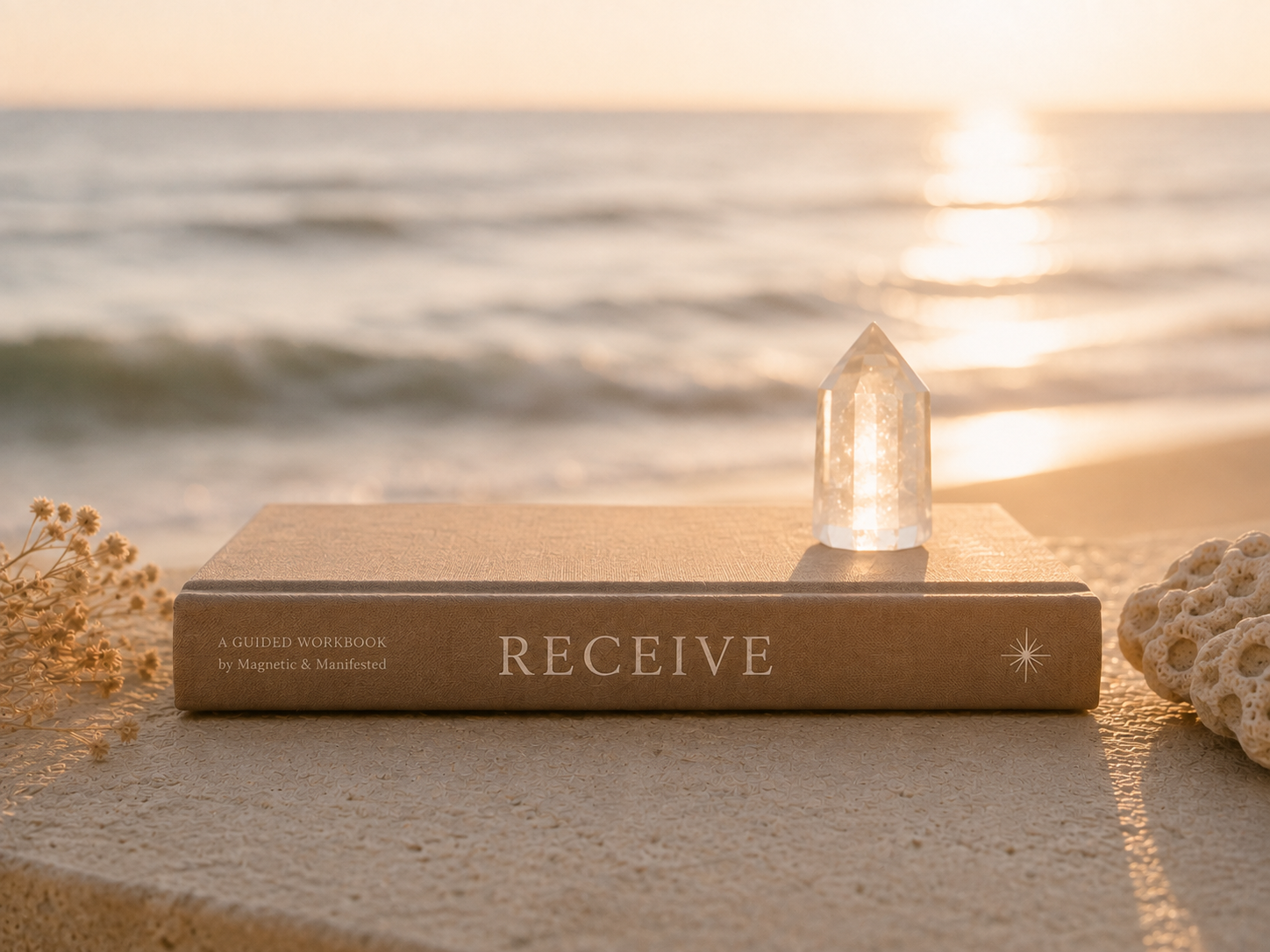 A book titled 'RECEIVE' placed on a sandy surface at the beach, with a crystal and decorative coral nearby and ocean in the background during sunset.