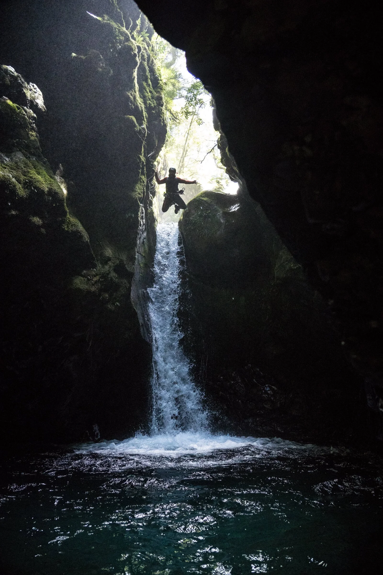 Person jumping in a narrow canyon with a waterfall and green vegetation, silhouetted against the light at the entrance of the canyon.