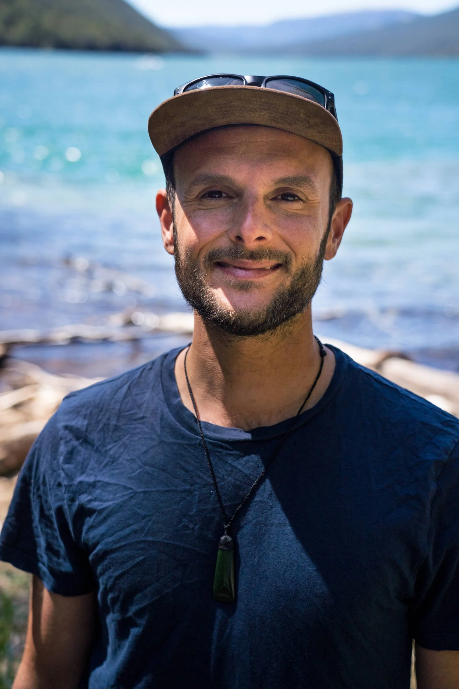 Adrien, wearing a brown cap with sunglasses on top, a blue shirt, and a necklace, smiling at the camera, with a background of water and mountains.