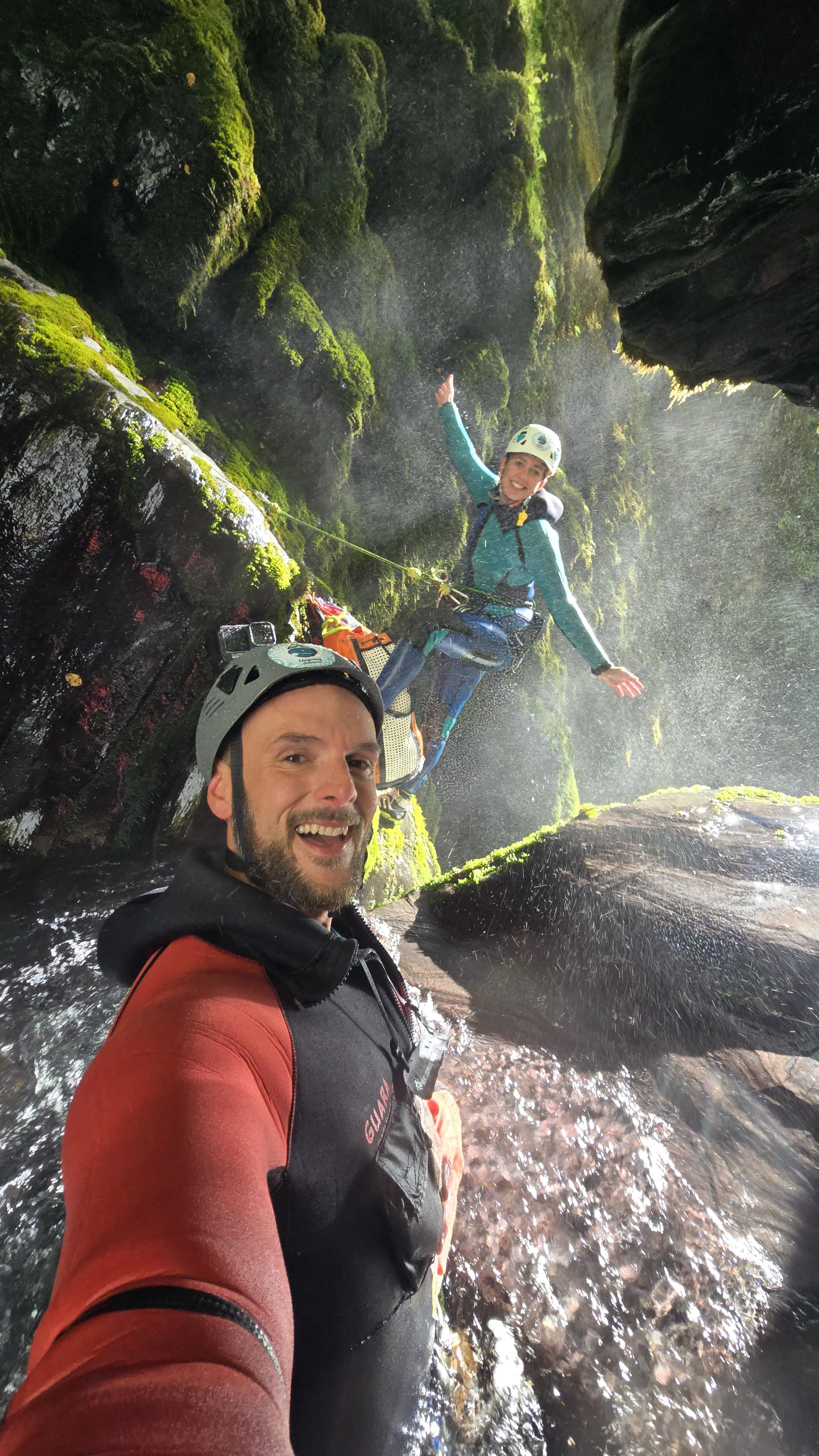 Two people in helmets and outdoor gear taking a selfie inside a mossy, rocky canyon with water flowing around them.