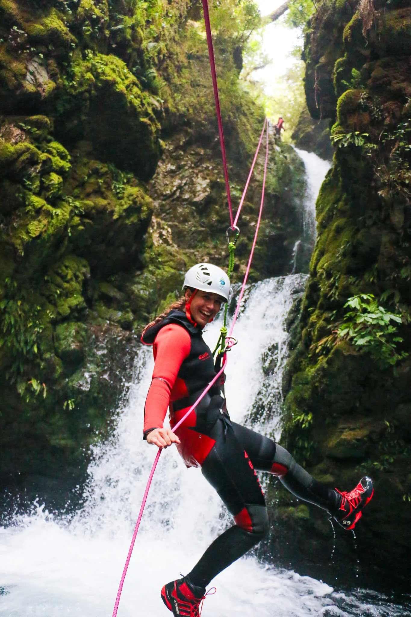 Person smiling while canyoning in a narrow rocky gorge with a waterfall, wearing a helmet, wetsuit, and climbing gear, holding a pink rope.