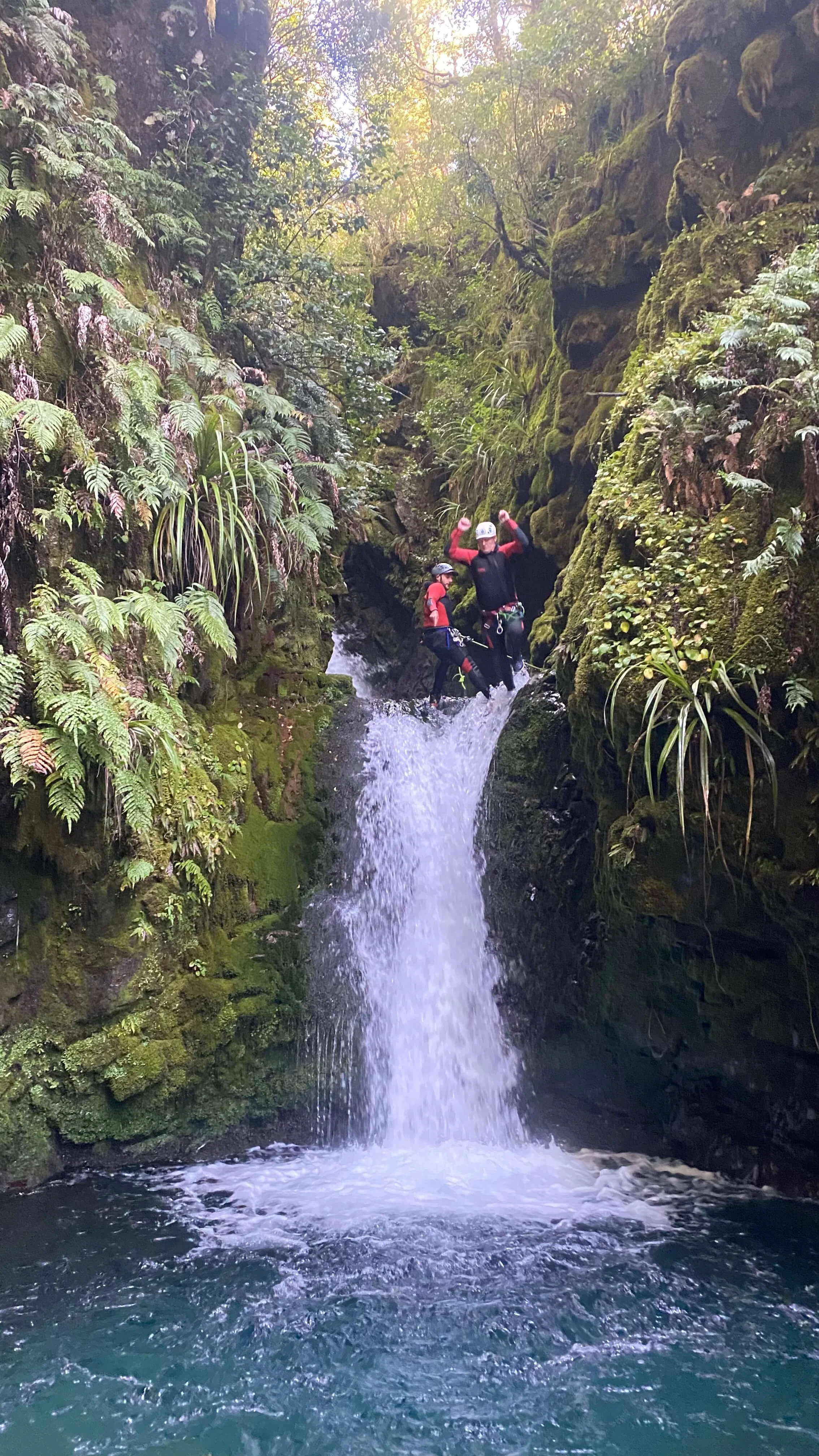 Two people in outdoor gear standing in a small waterfall in a lush forest canyon with moss-covered rocks and dense green foliage.