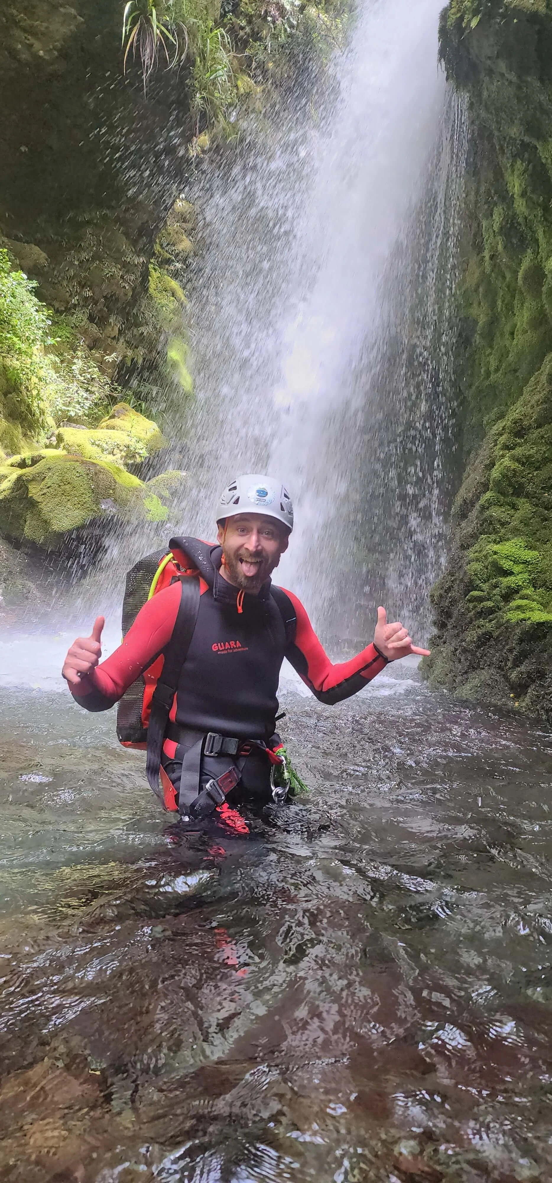 A man wearing a helmet and a black and red drysuit standing in a river near a waterfall, smiling and giving thumbs up.