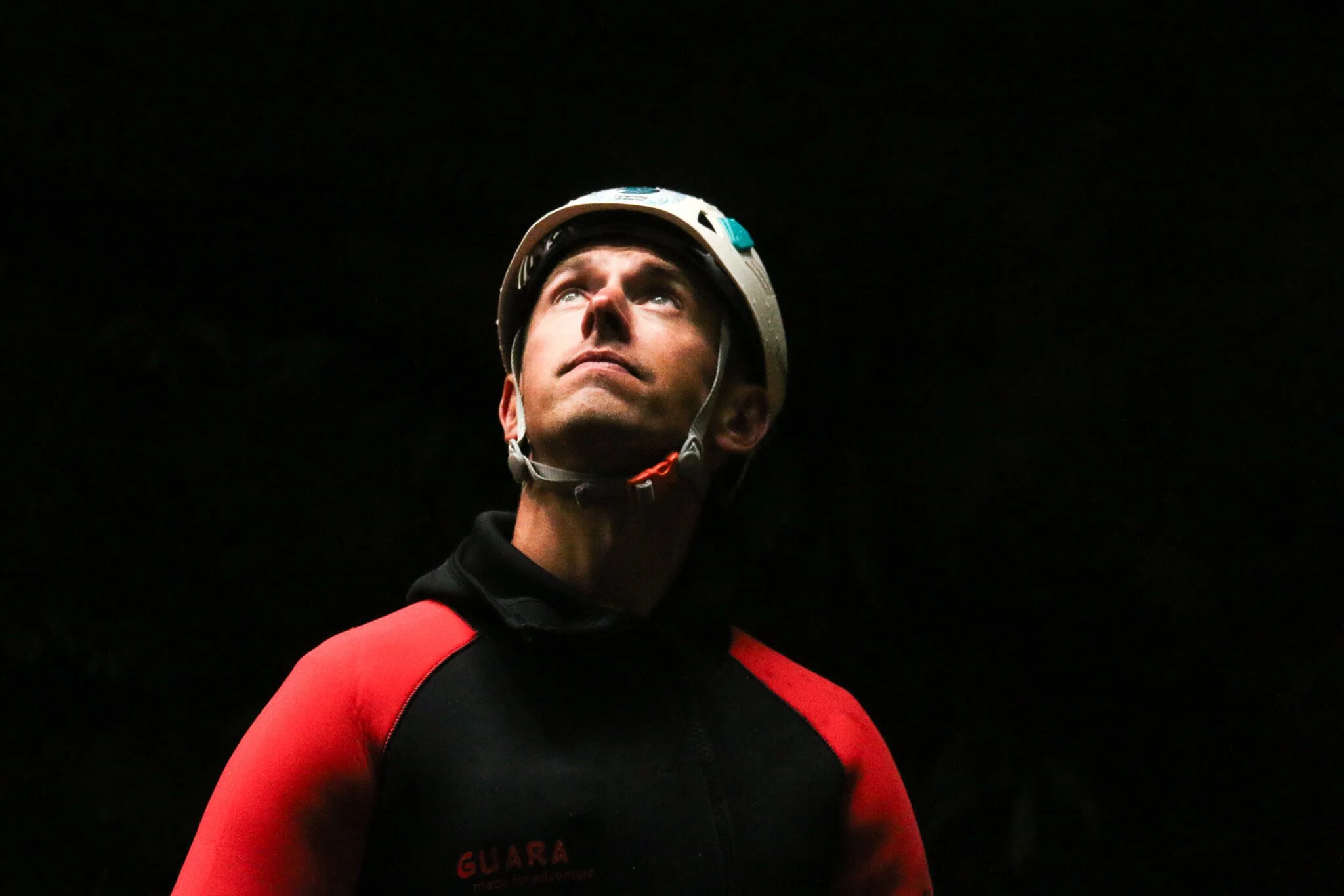 A person wearing a helmet and red and black athletic clothing, looking upward against a dark background.