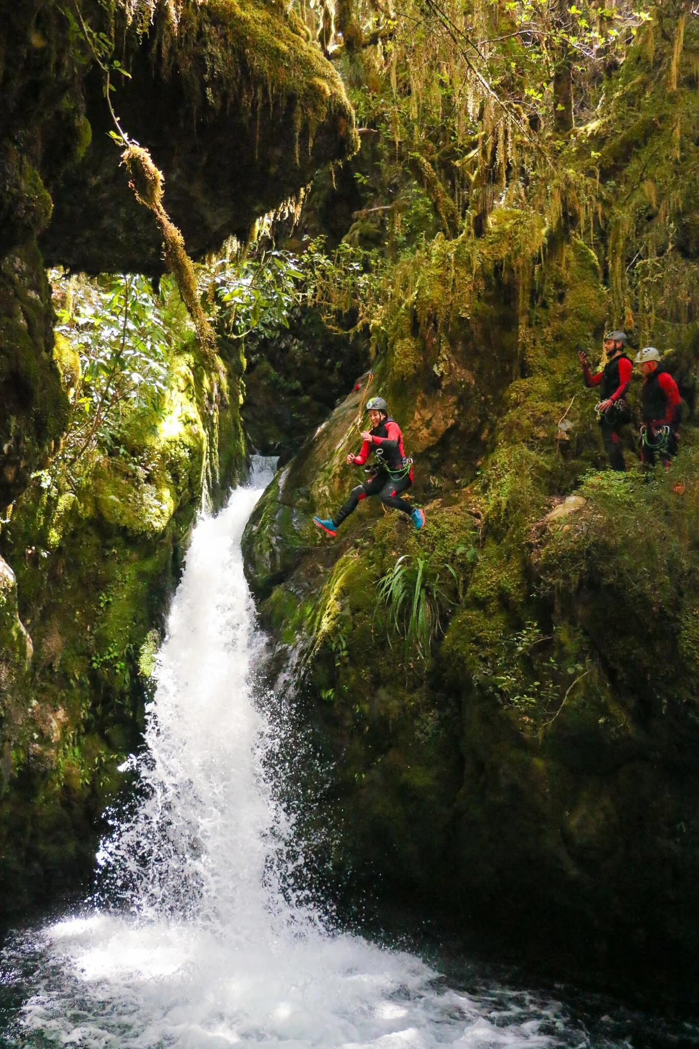 Group of people wearing helmets and wetsuits canyoning in a narrow, moss-covered rocky canyon with a waterfall.