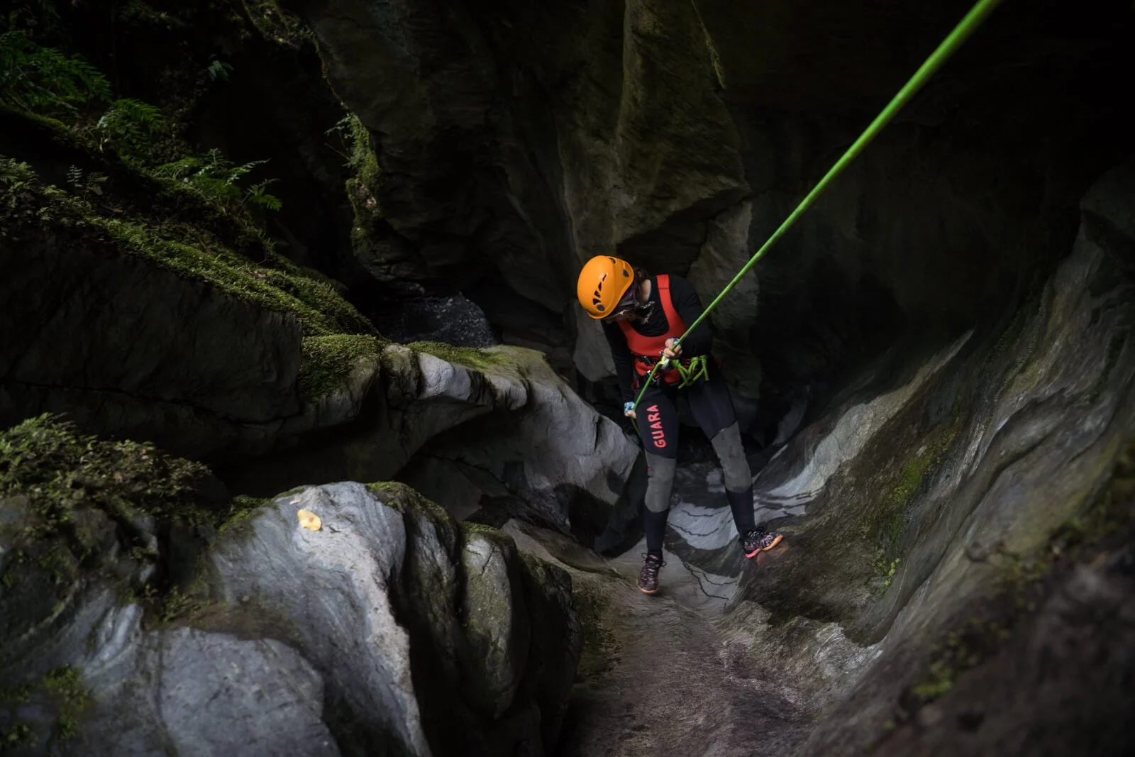A person wearing a yellow helmet, red safety harness, and black protective clothing is rappelling down a rocky canyon with green moss and vegetation.