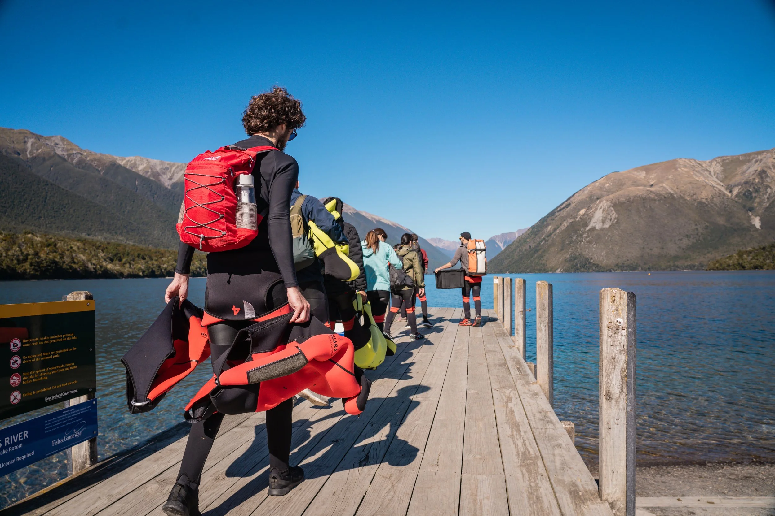 A group of people in outdoor gear walking on a wooden dock beside a lake, with mountains in the background under a clear blue sky.