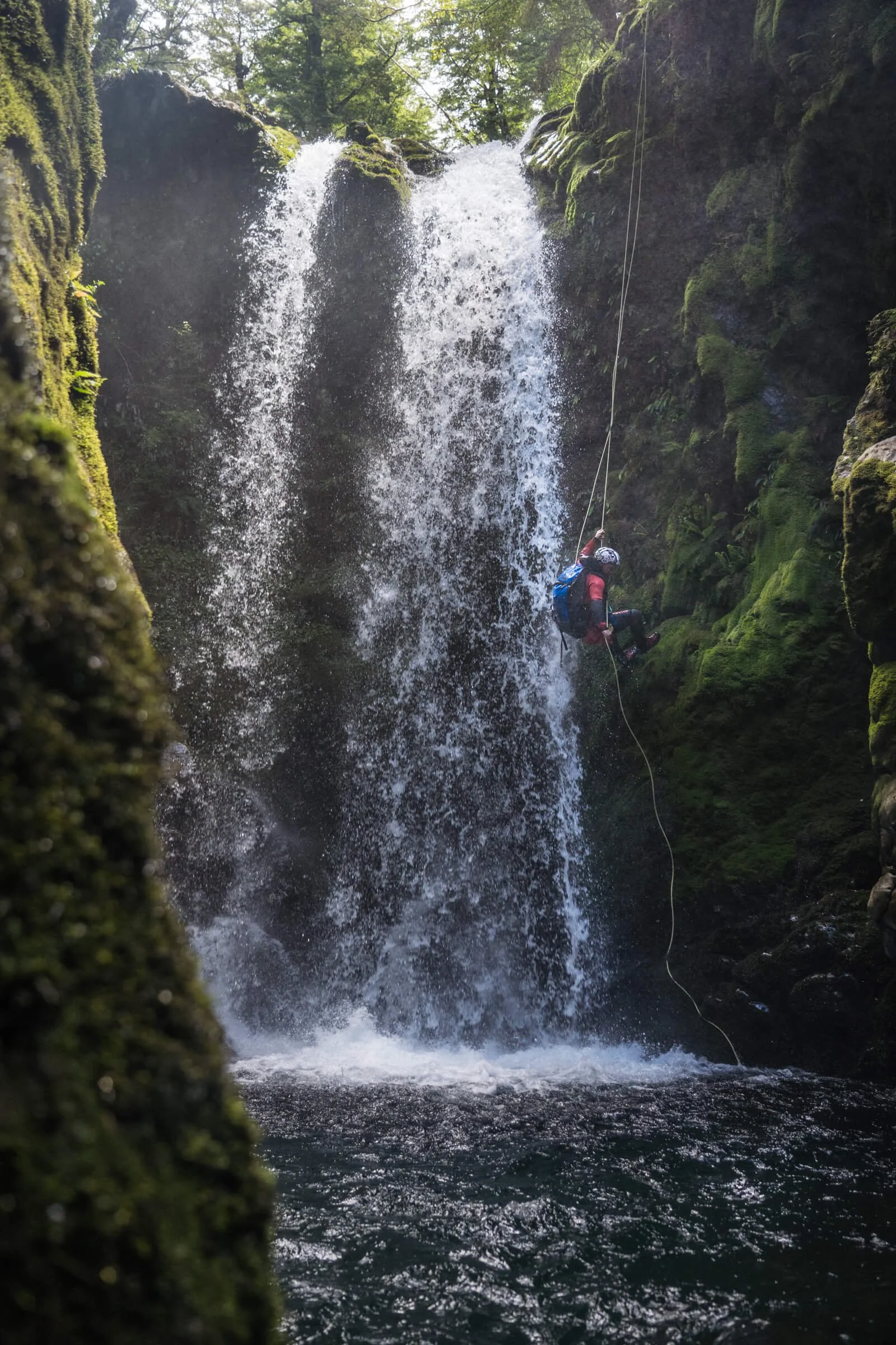 A person wearing a helmet and backpack is rappelling down a waterfall surrounded by moss-covered rocks and lush green trees.