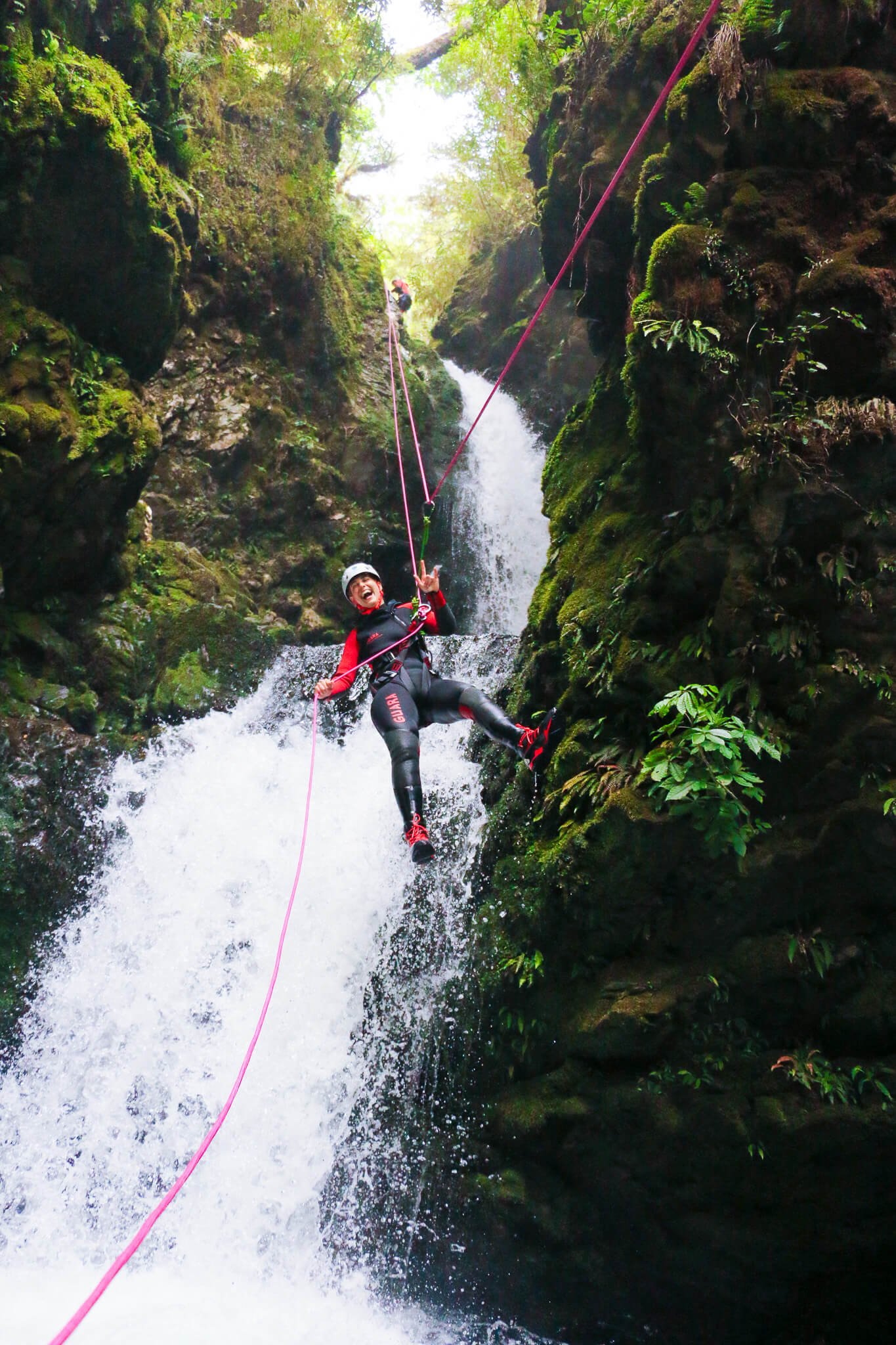 A person smiling and holding onto a rope while canyoning in a narrow, moss-covered rocky gorge with water rushing below.