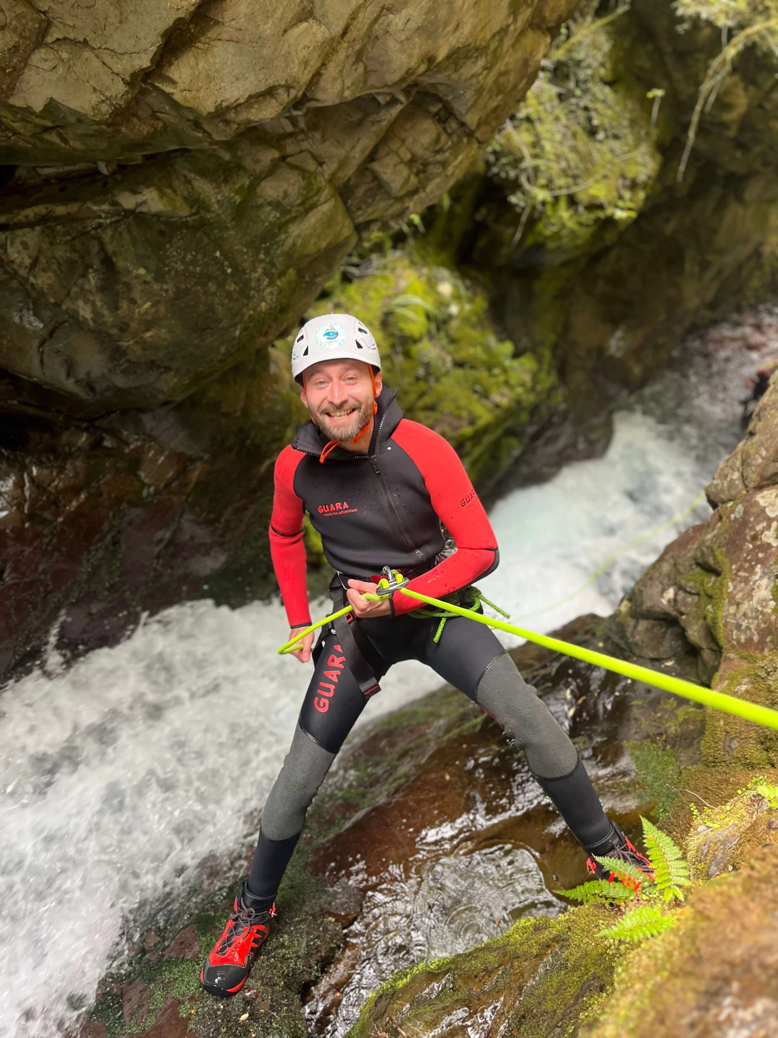 A man wearing a helmet and safety gear climbing on a rock near a rushing stream in a canyon or gorge.