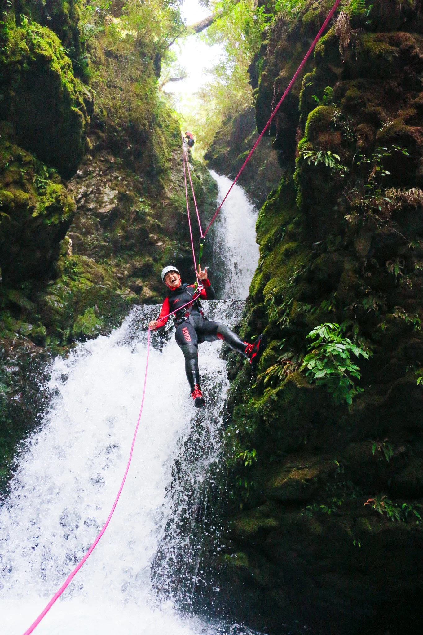 Two people rappelling down a waterfall in a lush green canyon. The person in the foreground is smiling, wearing a helmet, and holding onto a rope, while the person in the background is higher up on the rock face.